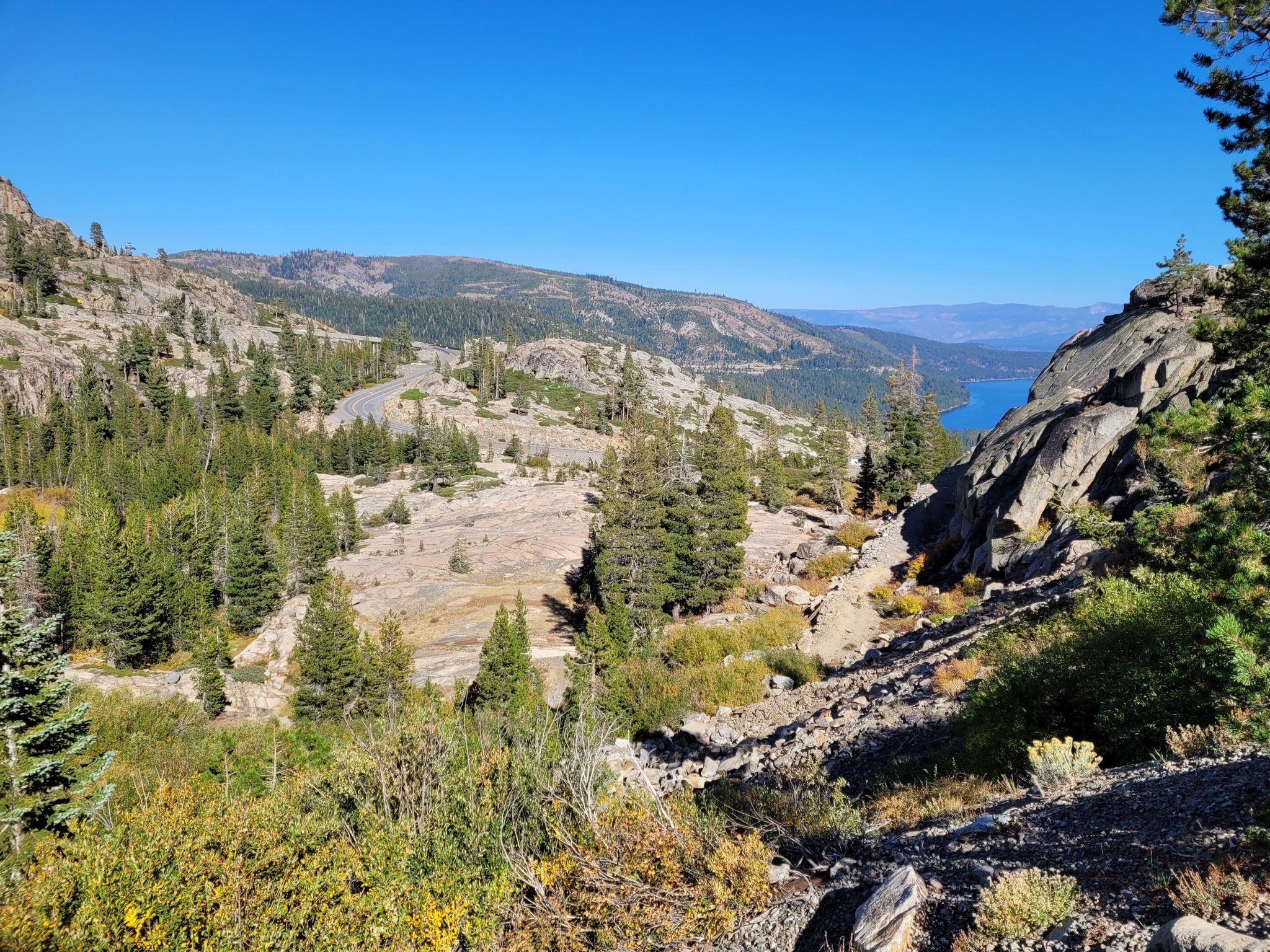 Sunny view of rocky, forested mountains with a winding road and distant lake. - Historic Donner Pass Trail