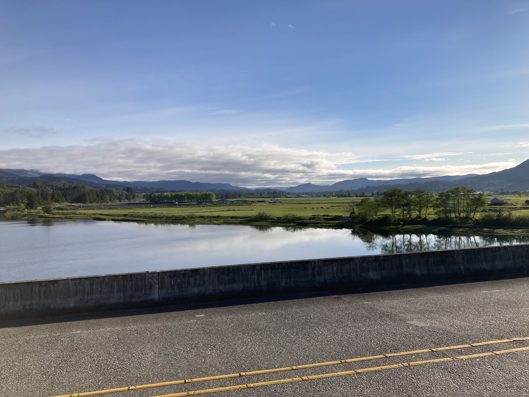 Roadside view of a calm river, green fields, and distant mountains under a cloudy sky. - Nehalem River – Nehalem River loop from Nehalem Bay State Park