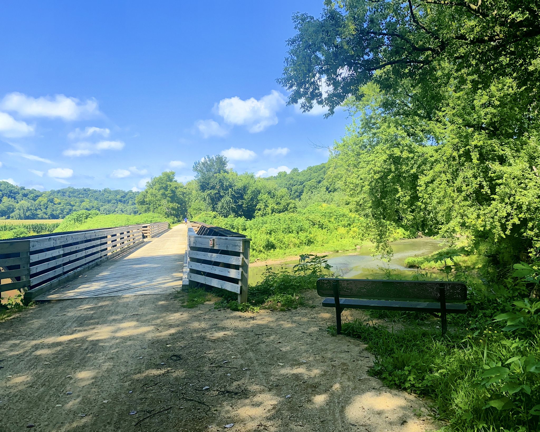 A wooden bridge and dirt path lead through a sunny, green landscape. - Heritage Trail
