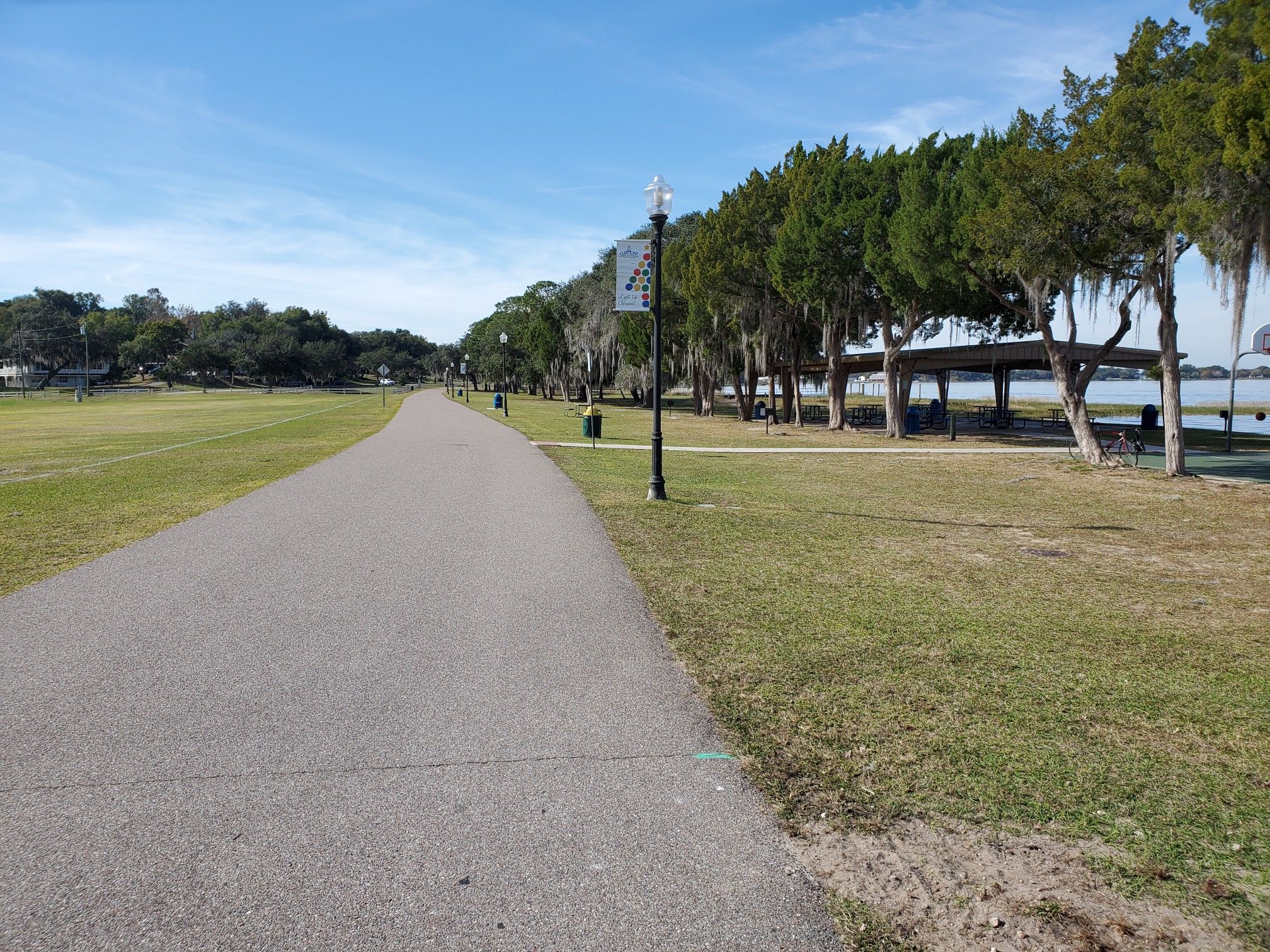 Paved path alongside a grassy field, trees, and lake on a sunny day. - Epic Cycles – Lake Hiawatha Preserve loop from Clermont