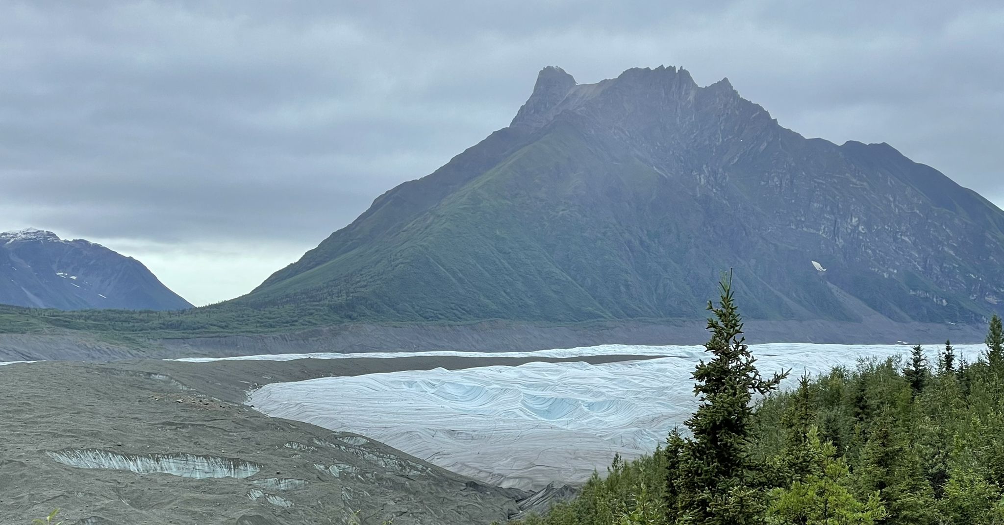 A large glacier with a towering mountain and green trees under an overcast sky. - Erie Mine Trail
