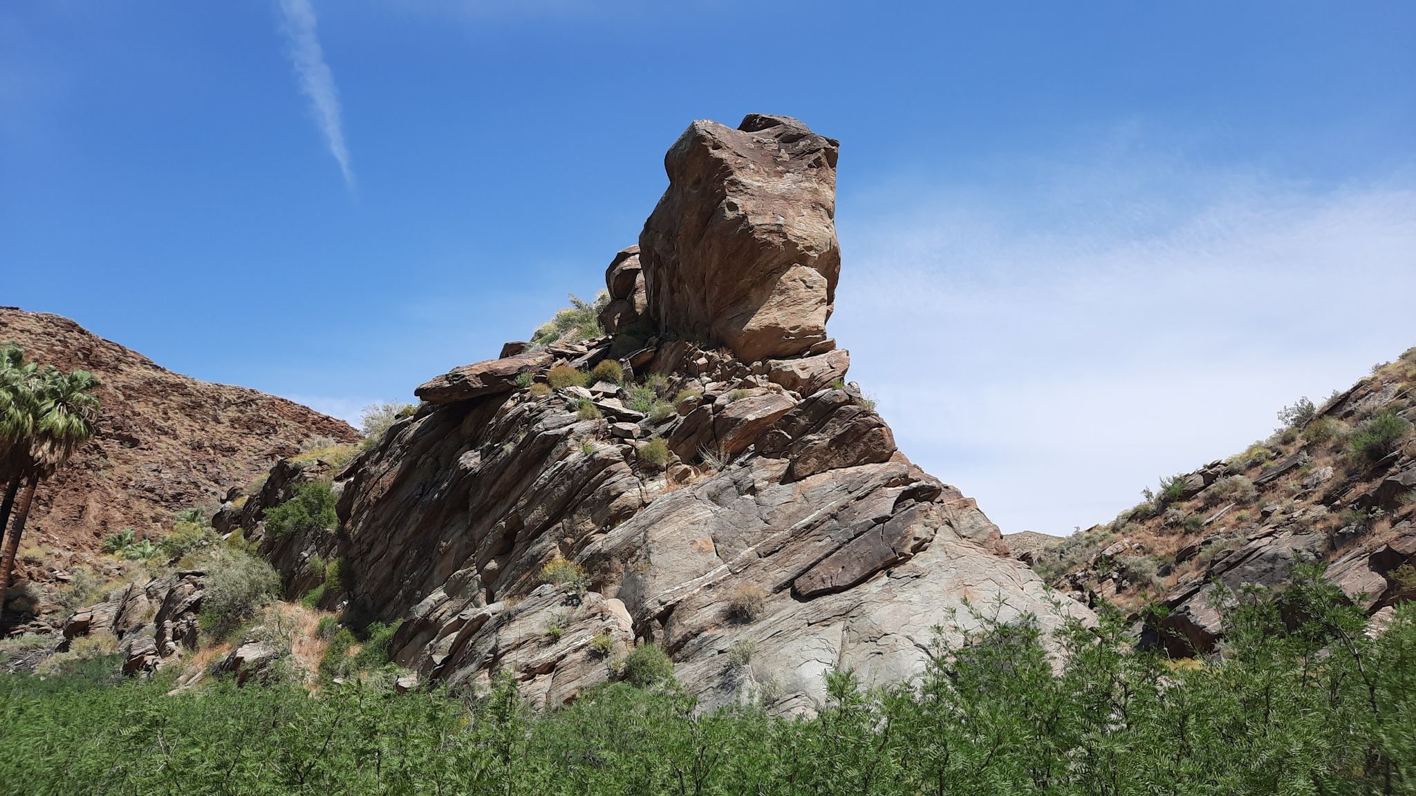 A towering, weathered rock formation stands in a sunny desert canyon with green shrubs. - Victor Trail Loop