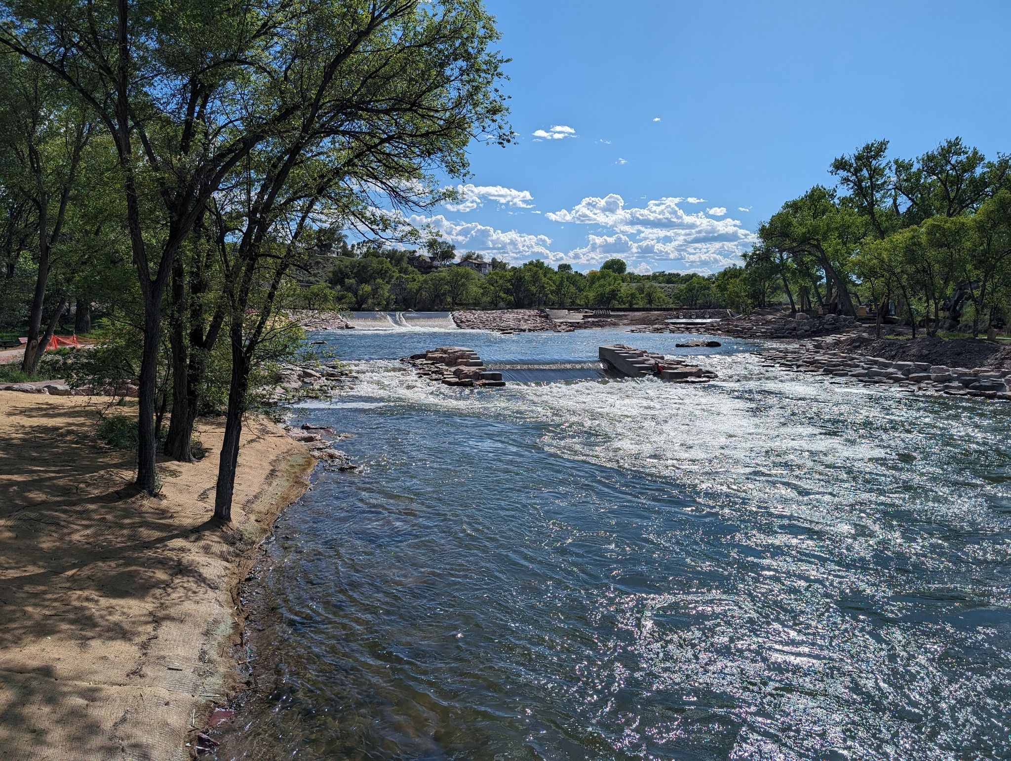 A wide river with rapids and a distant dam, surrounded by green trees. - The Arkansas River Trail loop from Pueblo