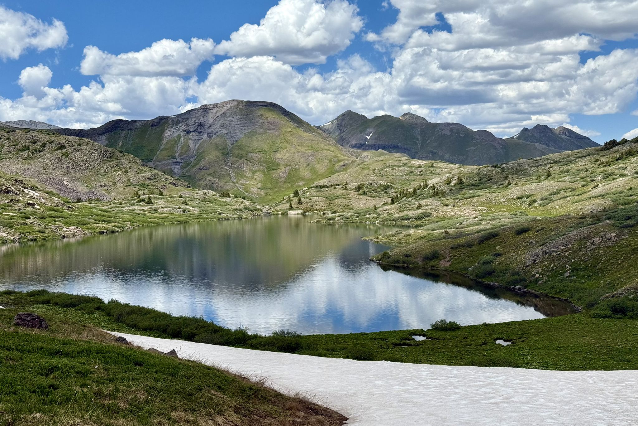 Mountain lake reflecting clouds, surrounded by green hills and a snow patch. - Highland Mary Lakes Loop Trail