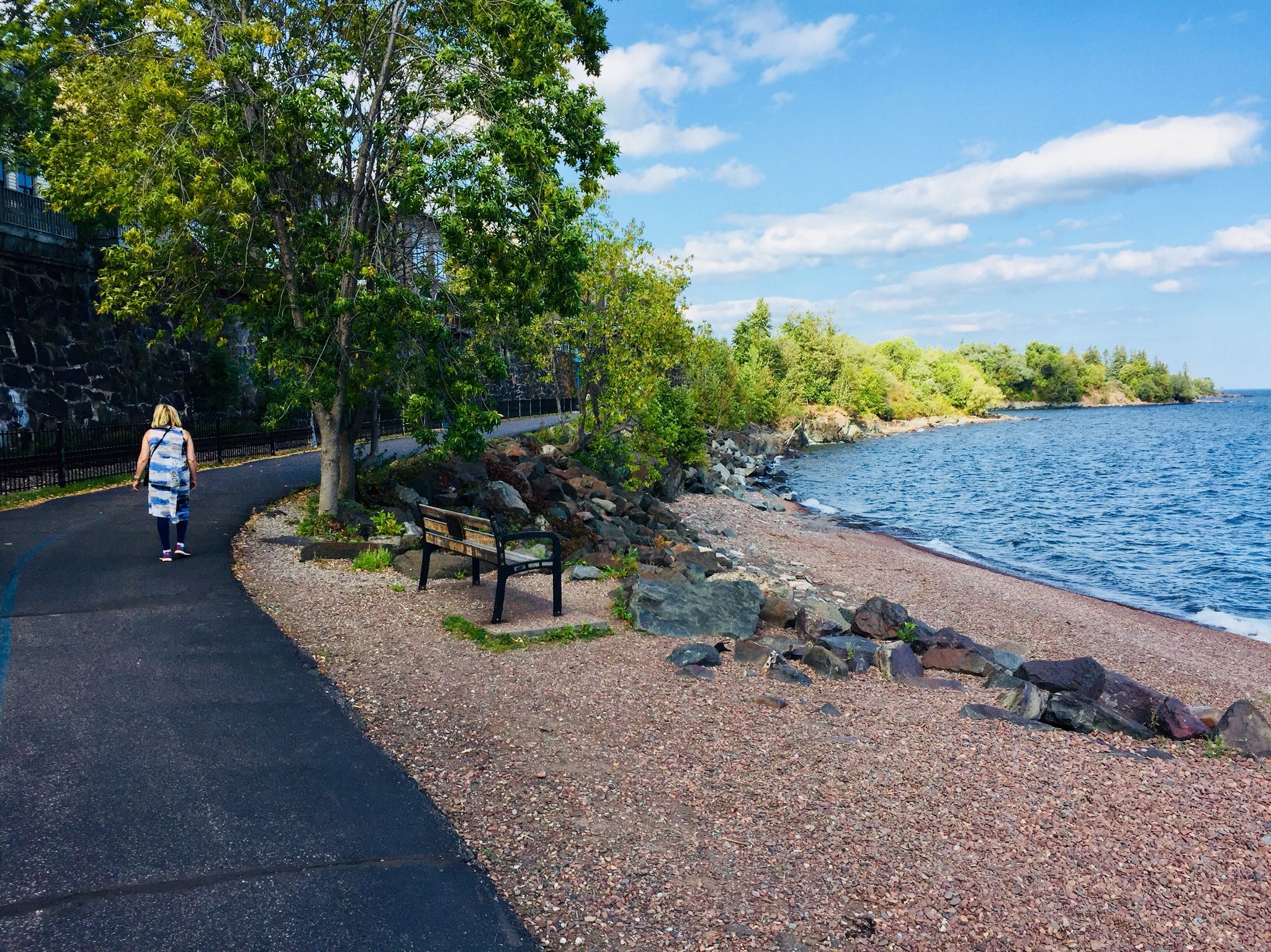 Person walks on a paved path next to a rocky beach and calm water. - Lakewalk loop from University of Minnesota Duluth