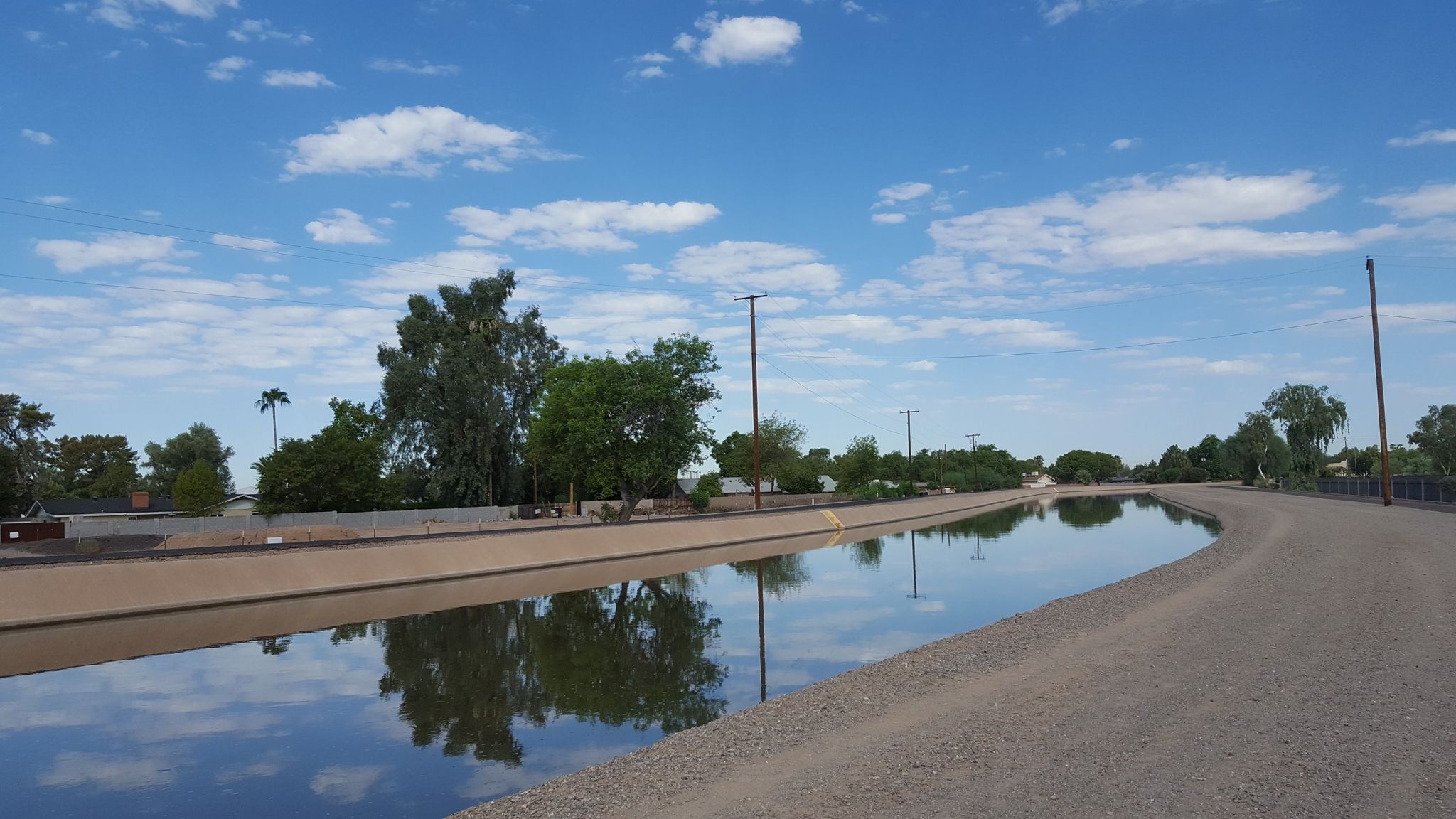 A wide canal reflecting a bright, cloudy sky, with dirt paths and trees. - Arizona Canal Trail: 32nd Street to 55th Ave