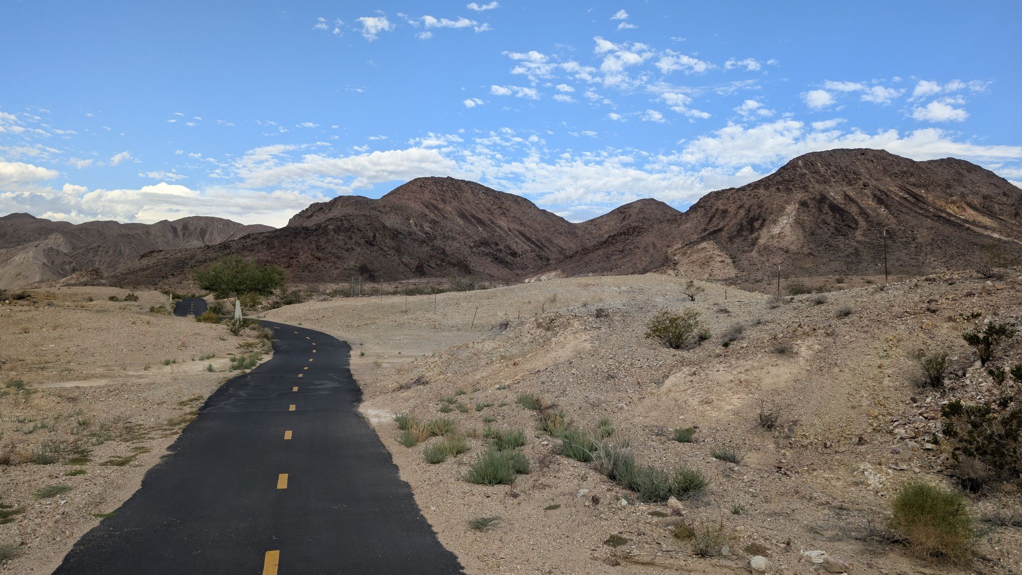 Paved road winding through a dry desert with mountains under a blue sky. - River Mountains Loop Trail