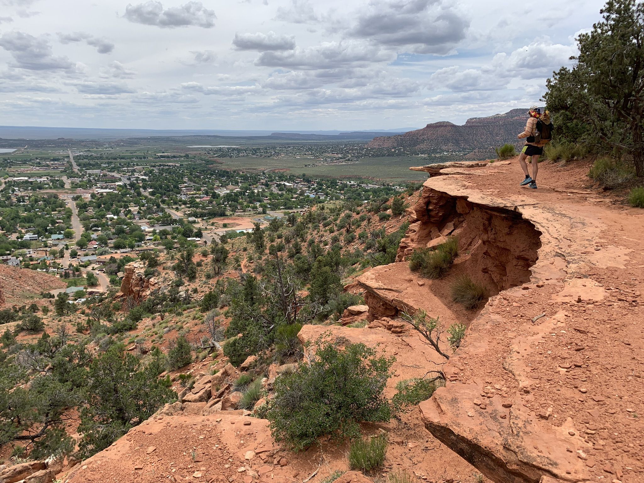 Person stands on a red rock cliff overlooking a distant valley town. - Kanab City Trail, Cliffs Trail, and Toms Canyon Loop
