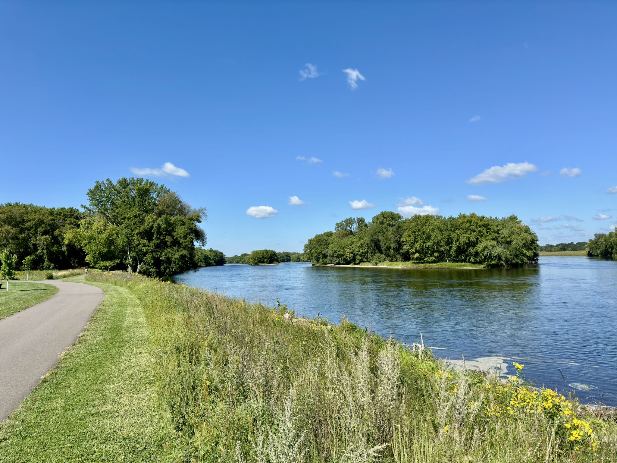 Paved path next to a wide river with forested islands under a sunny sky. - Otsego County Park Walking Path