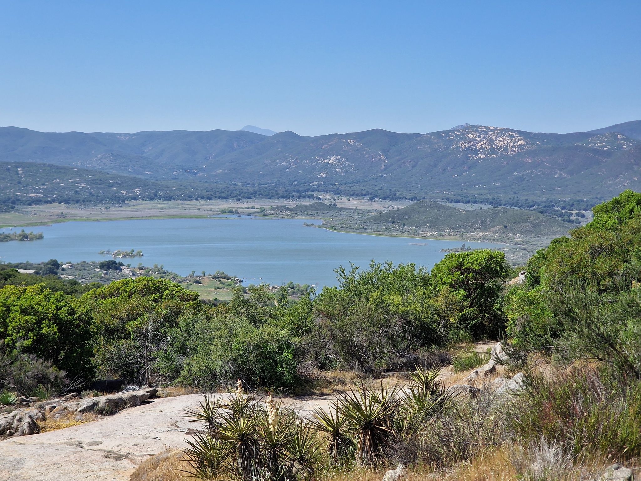 A serene lake surrounded by green hills and distant mountains under bright sun. - Morena Butte loop from Lake Morena Drive & Lake Morena Campground