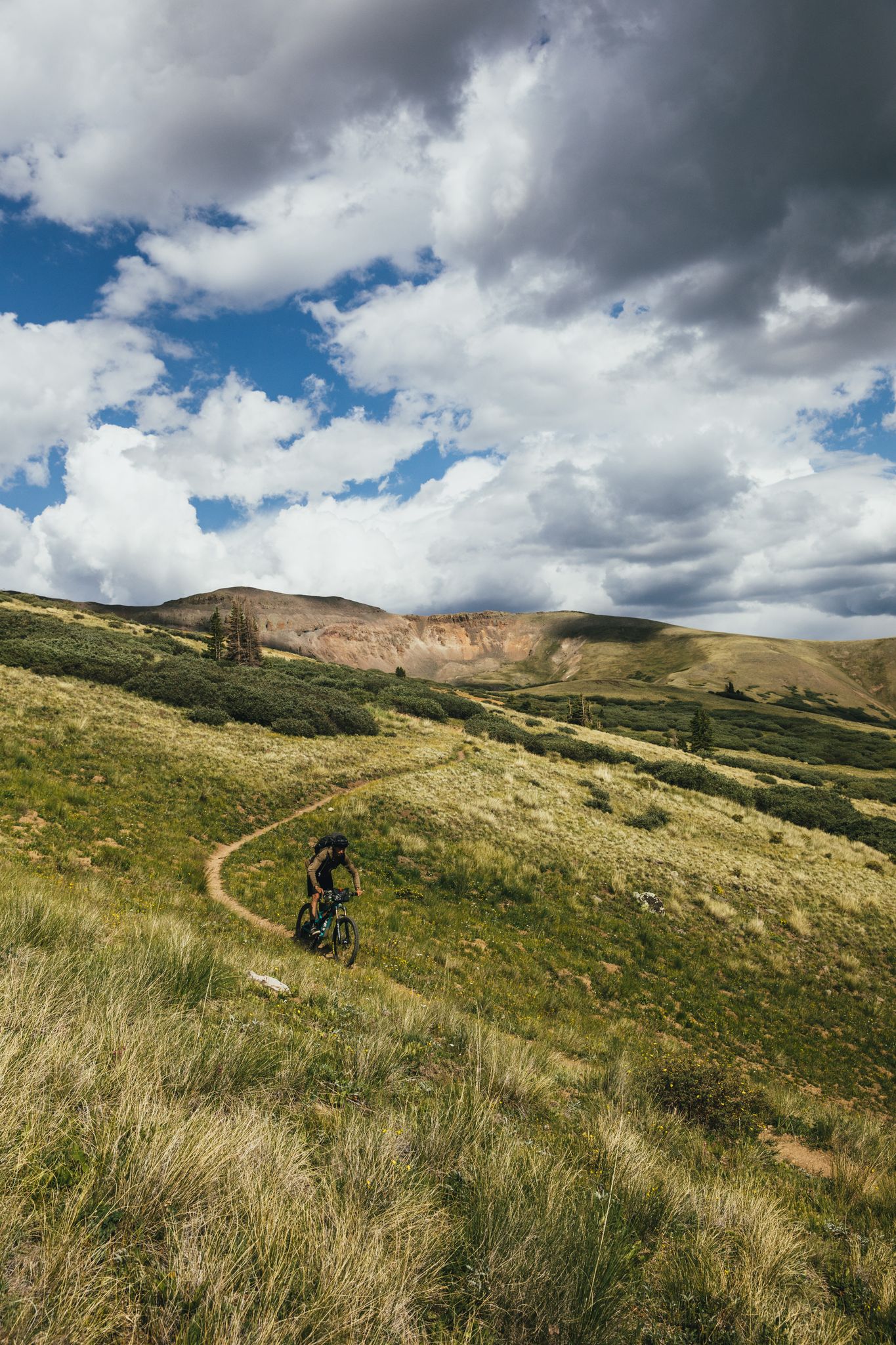 Mountain biker on a winding trail up a grassy hillside under cloudy sky. - Colorado Trail: Segment 22