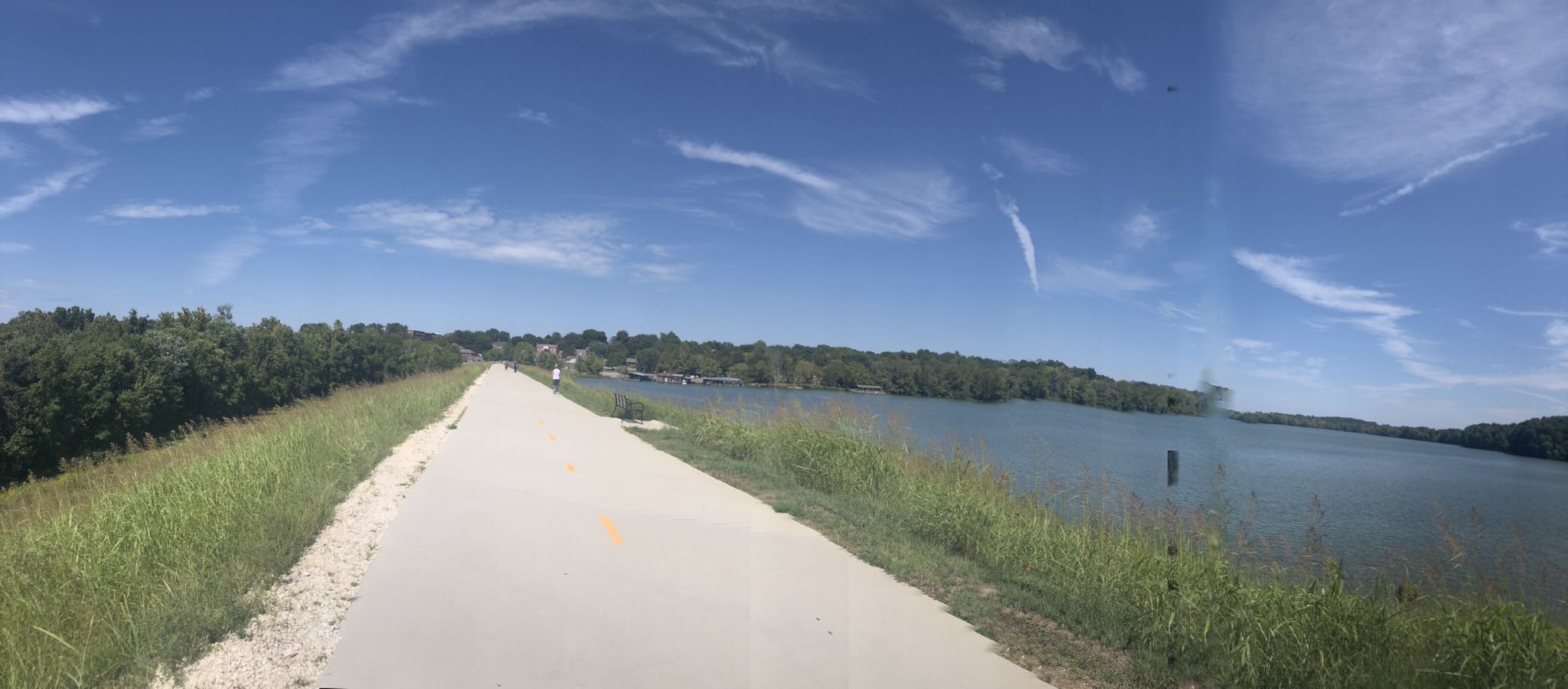 A wide paved path runs alongside a large lake under a blue sky. - Lake Fayetteville Trail – Boardwalk along Spring Creek loop from Johnson