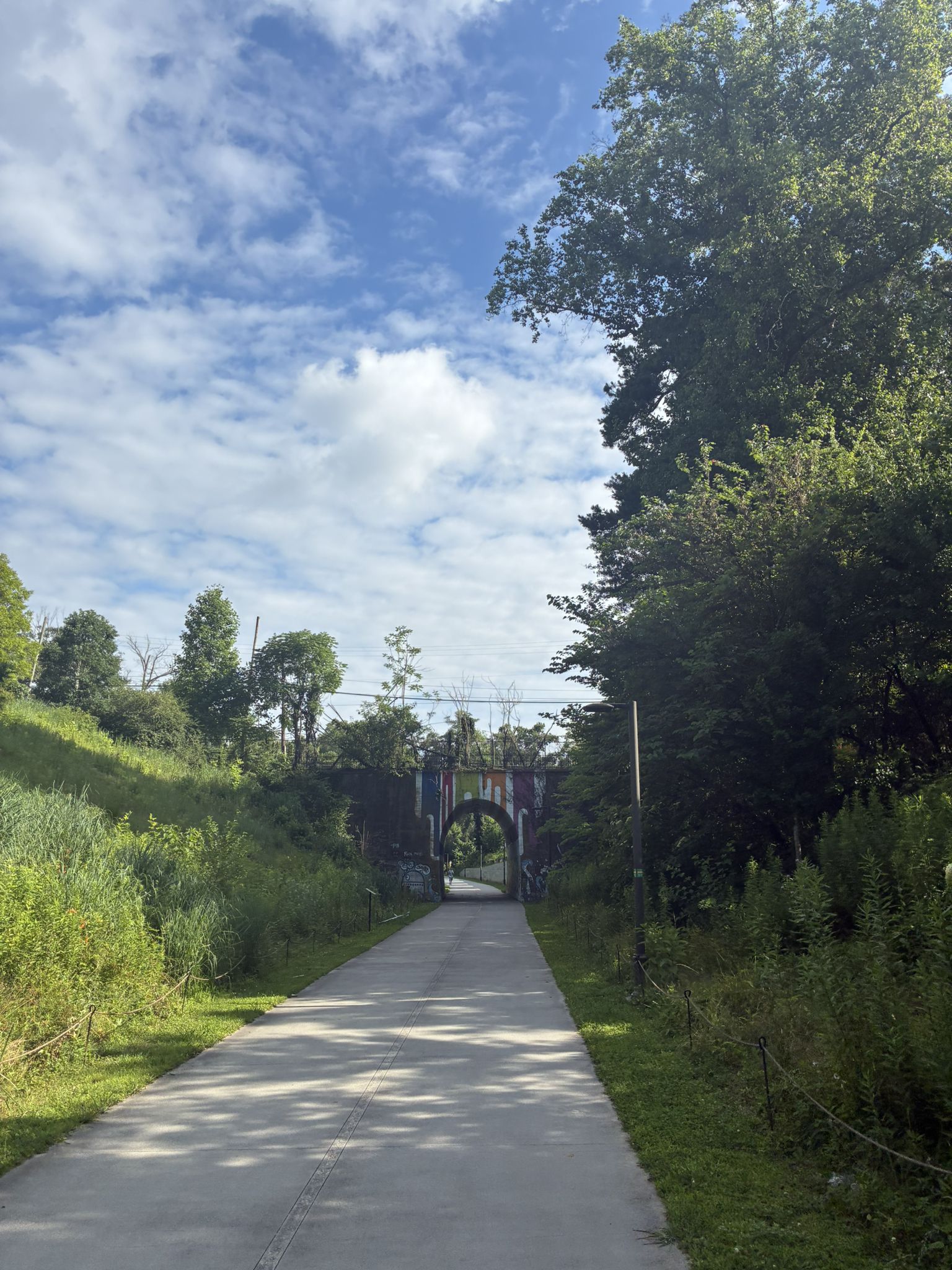 A paved path leads towards a dark, arched tunnel under a partly cloudy sky. - Atlanta Beltline Trail