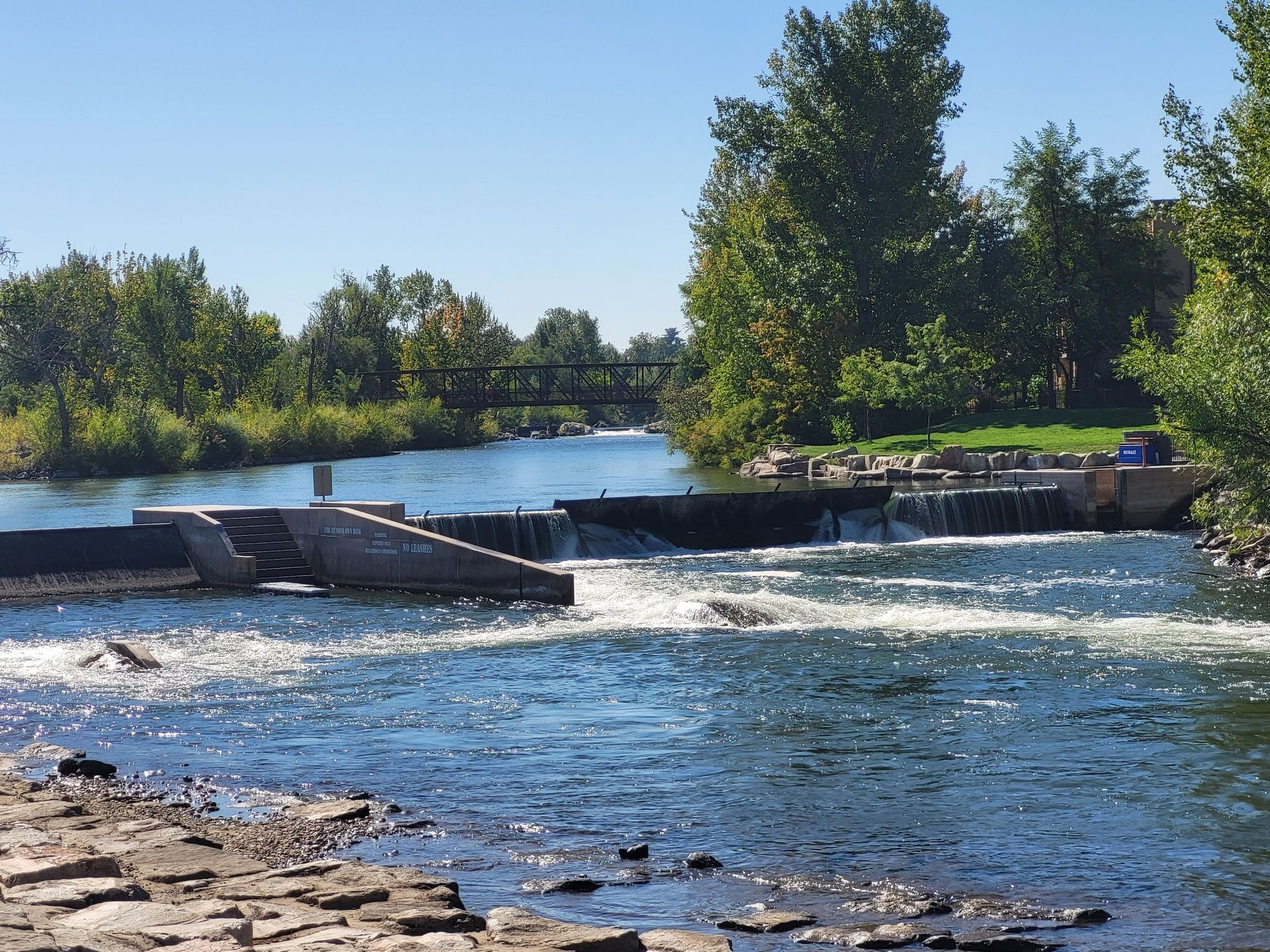 Wide river with a dam, surrounded by green trees on a sunny day. - Boise River Greenbelt: Garden City to Eagle Road