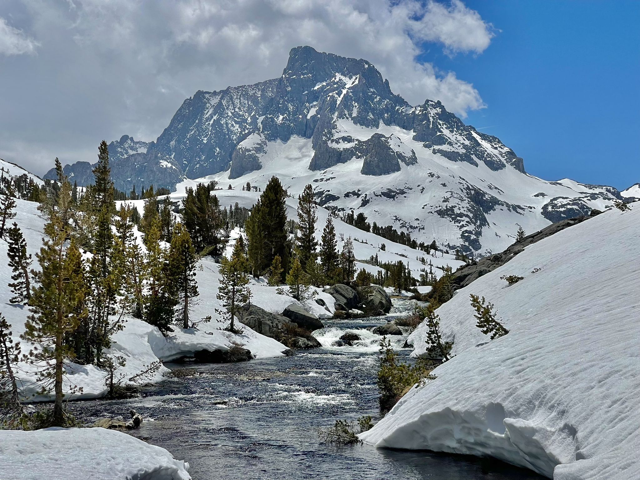 Rushing river through snowy banks and evergreen trees, with a majestic snow-capped mountain. - Thousand Island Lake via Rush Creek Trail