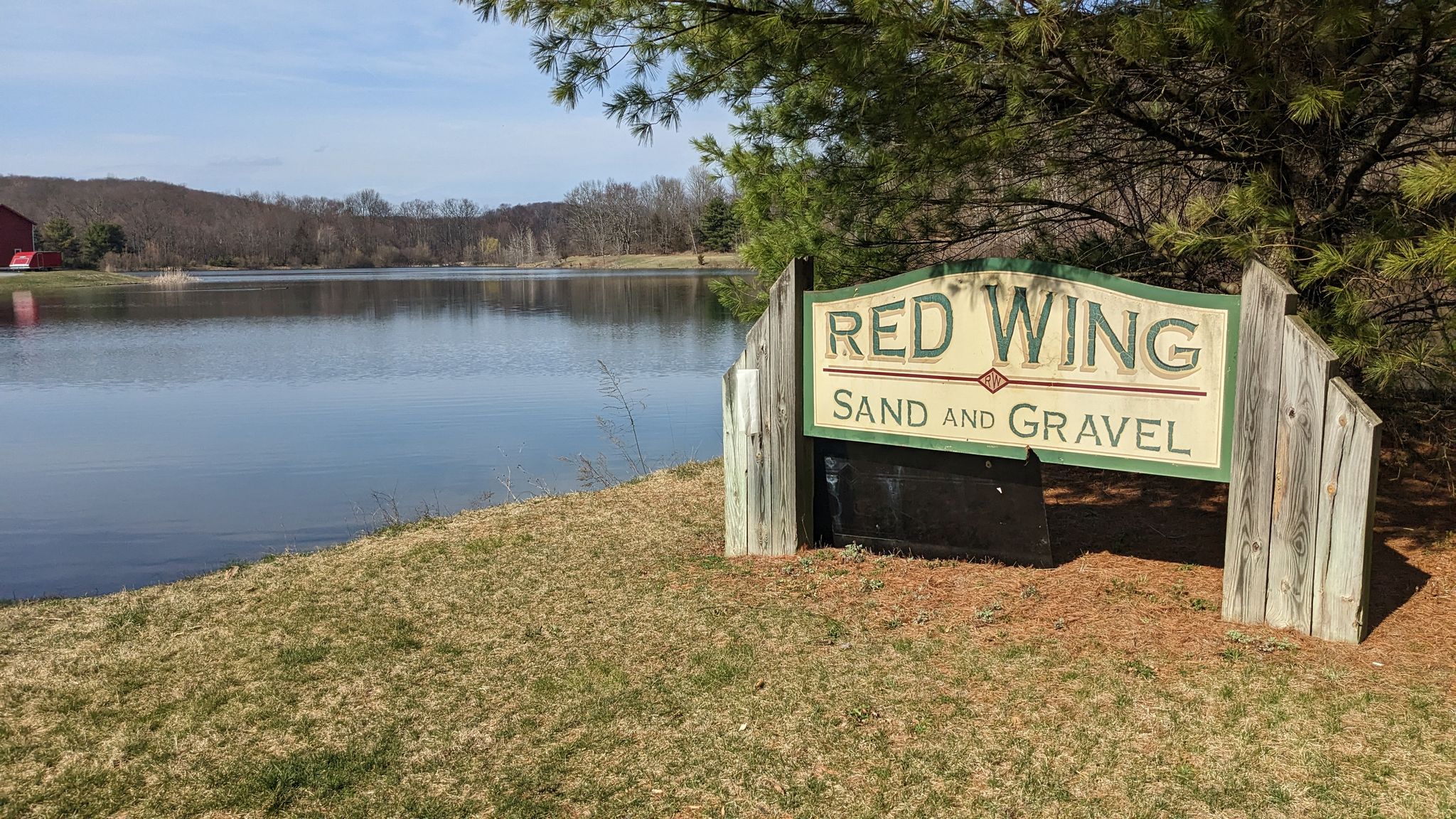 Wooden sign on grassy bank beside a calm lake under a bright sky. - Red Wing Lakes Loop