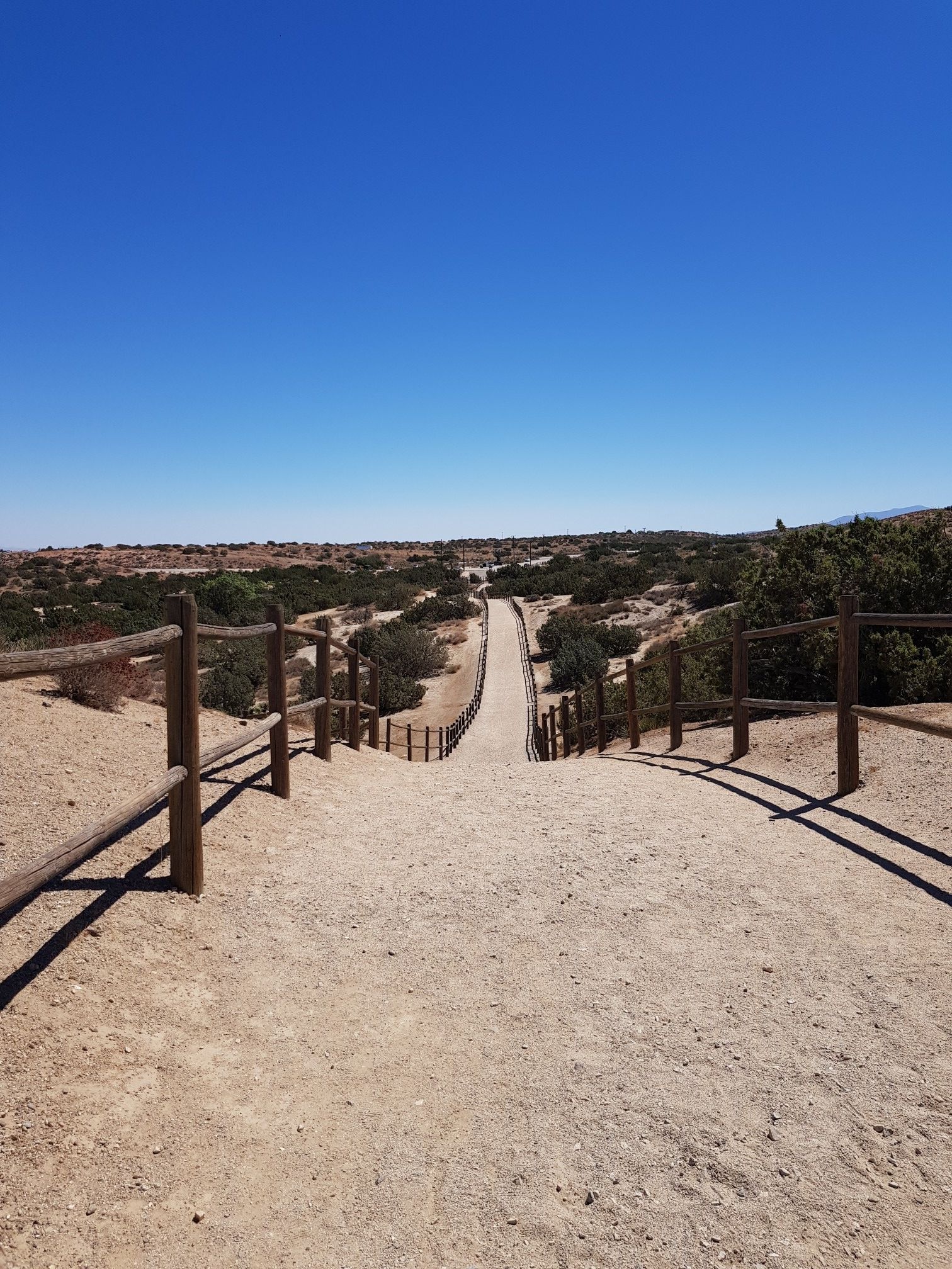 A dirt path slopes down between wooden fences into a distant valley. - Barrel Springs Trail