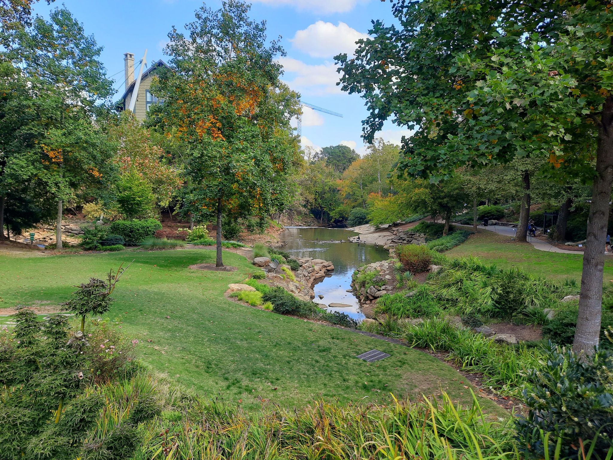 Lush green park with a winding stream, trees, and a distant building under blue sky. - Paris Mountain and Swamp Rabbit Trail Loop