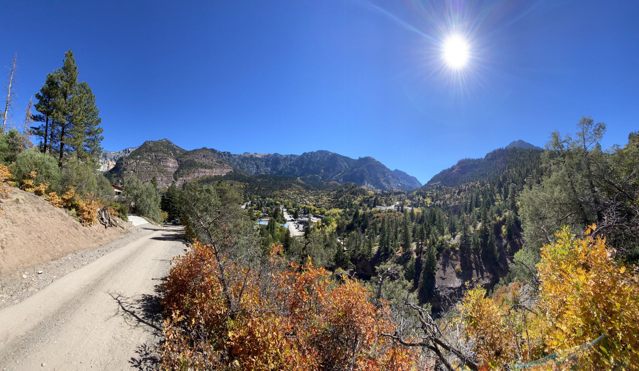 A dirt road overlooks a mountain valley with autumn foliage under bright sun. - Ouray Perimeter Trail