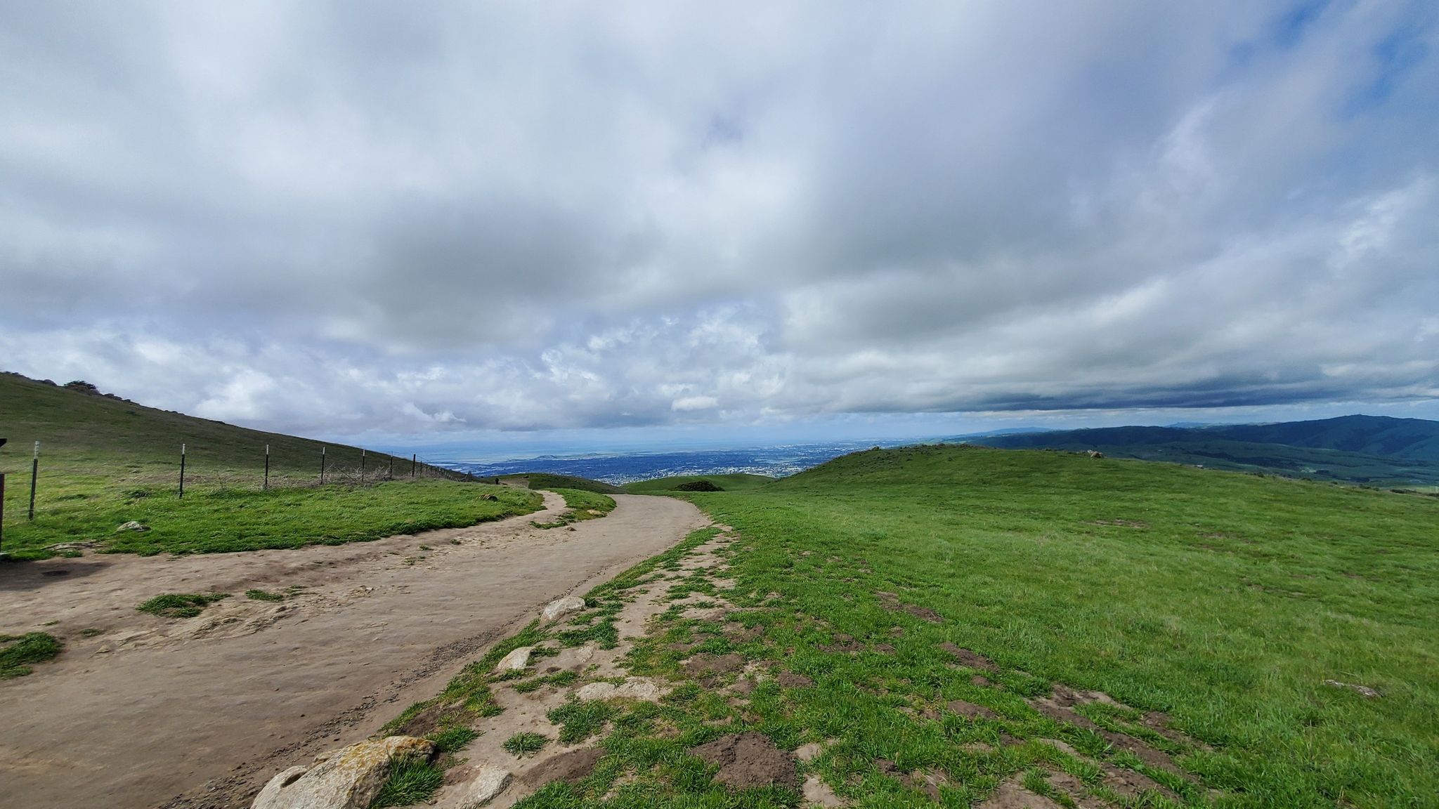 A dirt path curves through vibrant green hills under a dramatic cloudy sky. - Mission Peak via Hidden Valley Trail