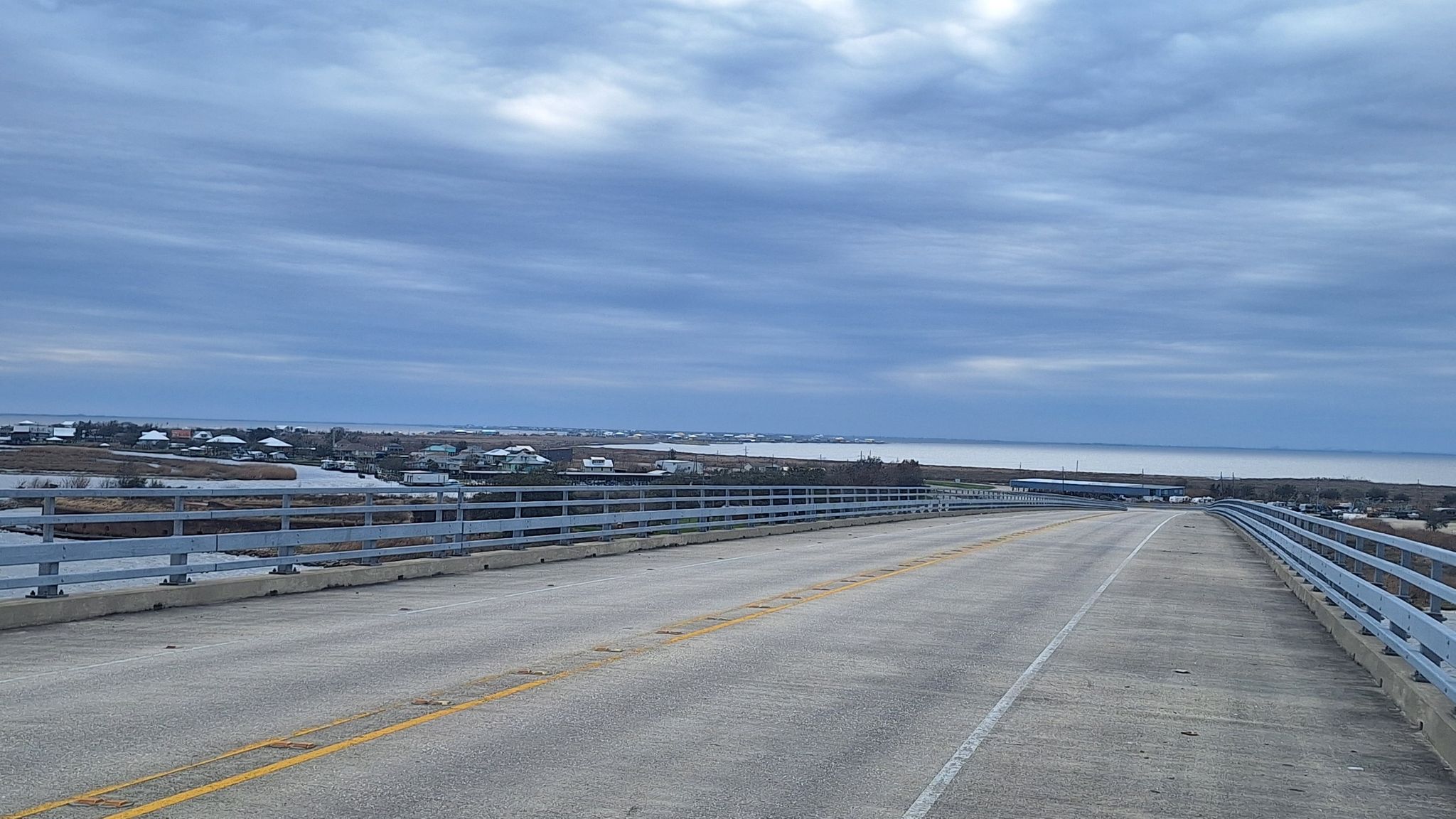 Empty bridge extends over calm water towards distant coastal homes under cloudy sky. - Chef Menteur Highway to West Pearl Bridge