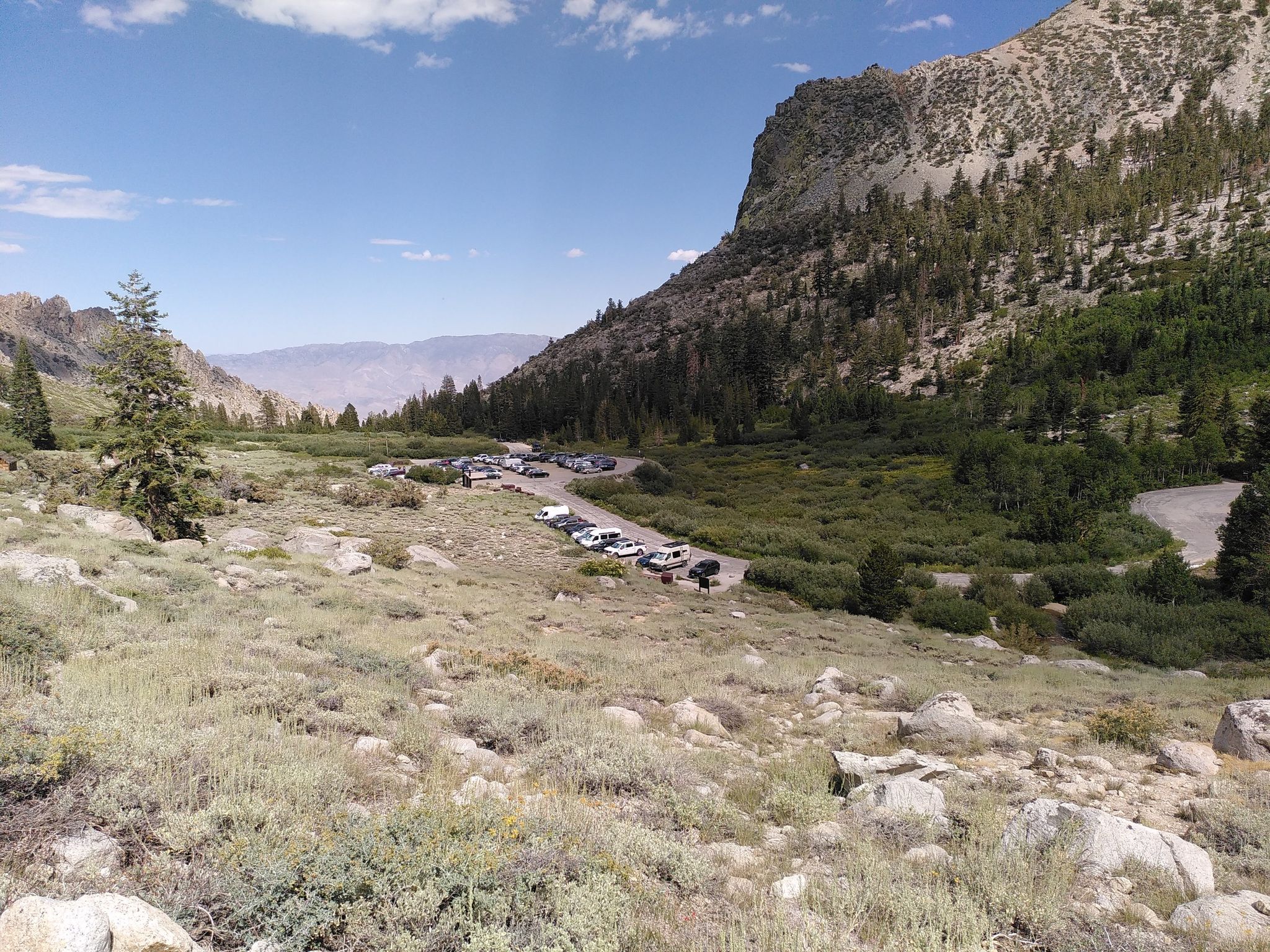 Parking lot in a mountain valley surrounded by green hills and distant peaks. - Little Pothole Lake loop from Kearsarge Peak