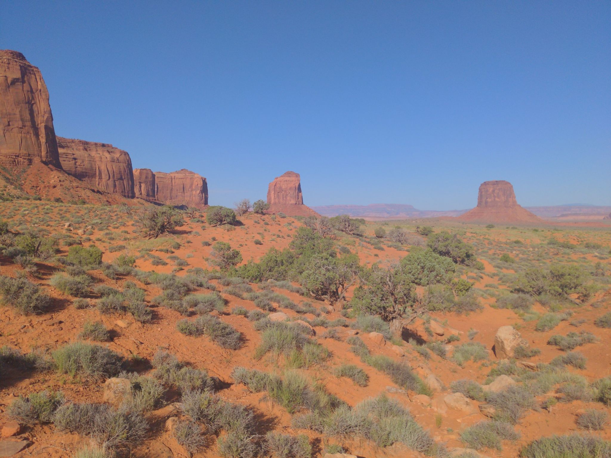 Vast desert landscape with red rock mesas and sparse green shrubs under clear sky. - Monument Valley Campground Lee Cly Trail