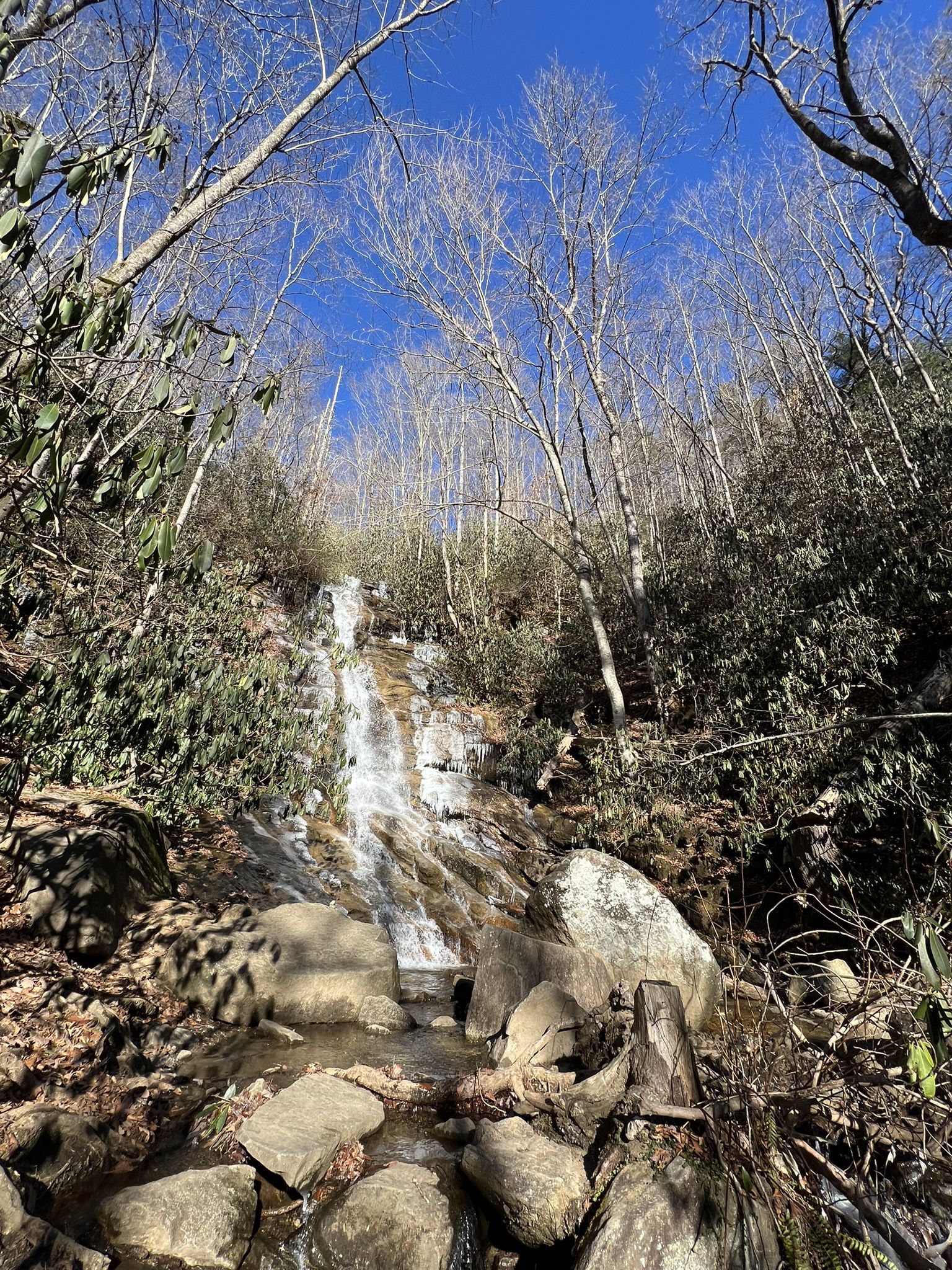 Tall, narrow waterfall cascades down a rocky, forested slope under a clear blue sky. - Jones Gap Falls Trail