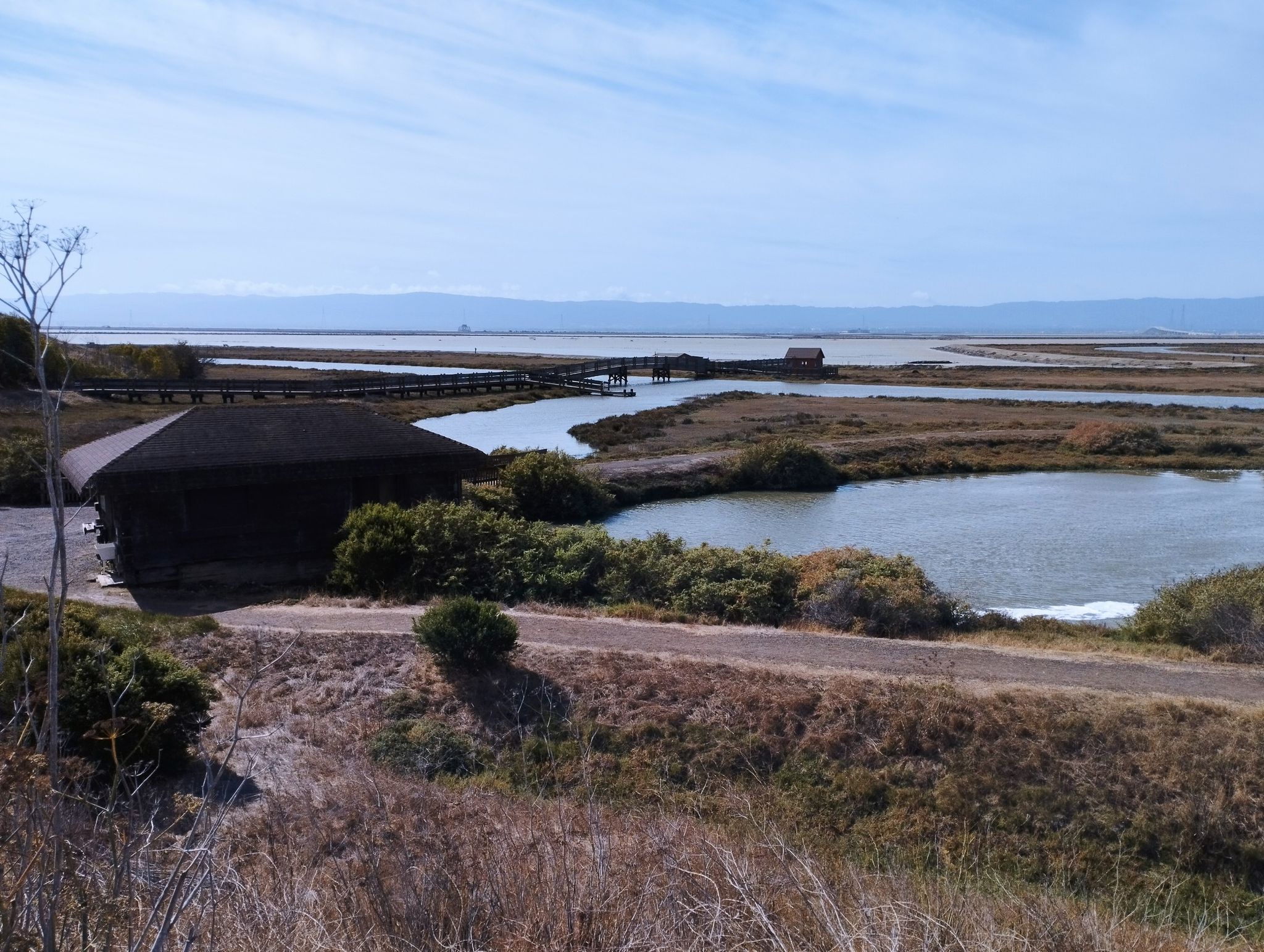 A dark building by calm marsh water with a distant boardwalk under clear skies. - San Francisco Bay Trail: Dumbarton Bridge Loop