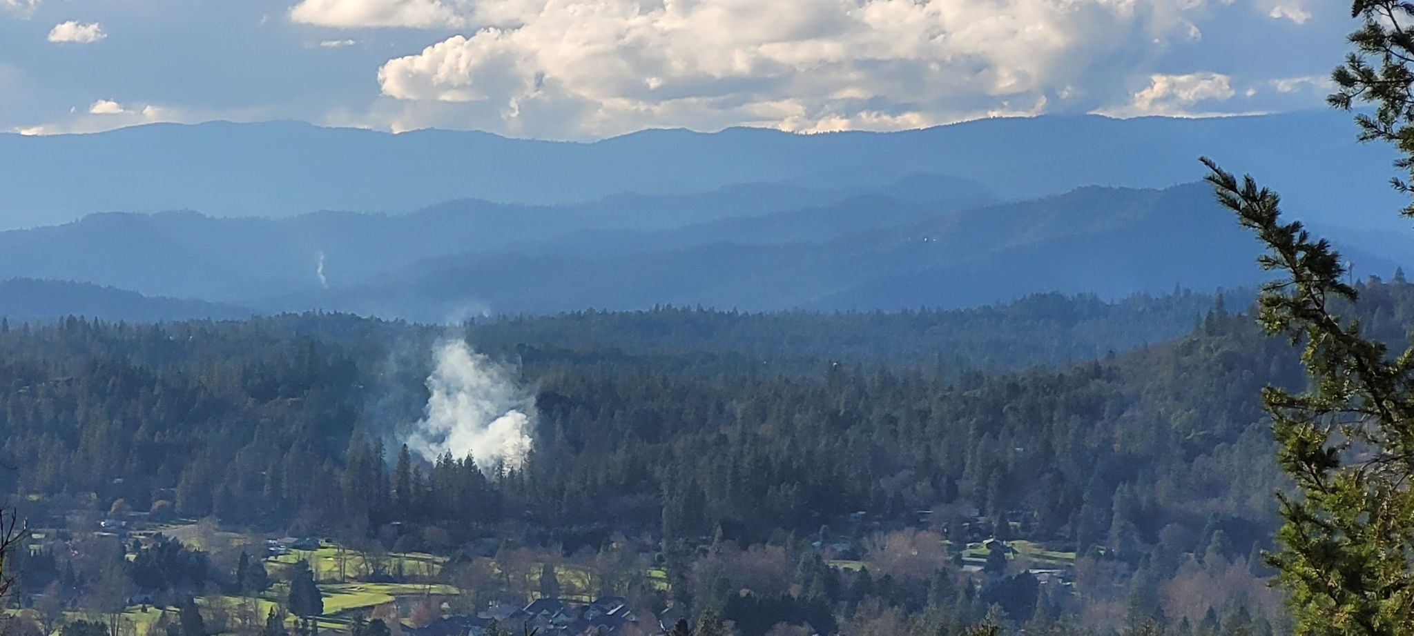 Smoke plume rising from a forested valley with distant mountains under clouds. - Outback Loop Trail