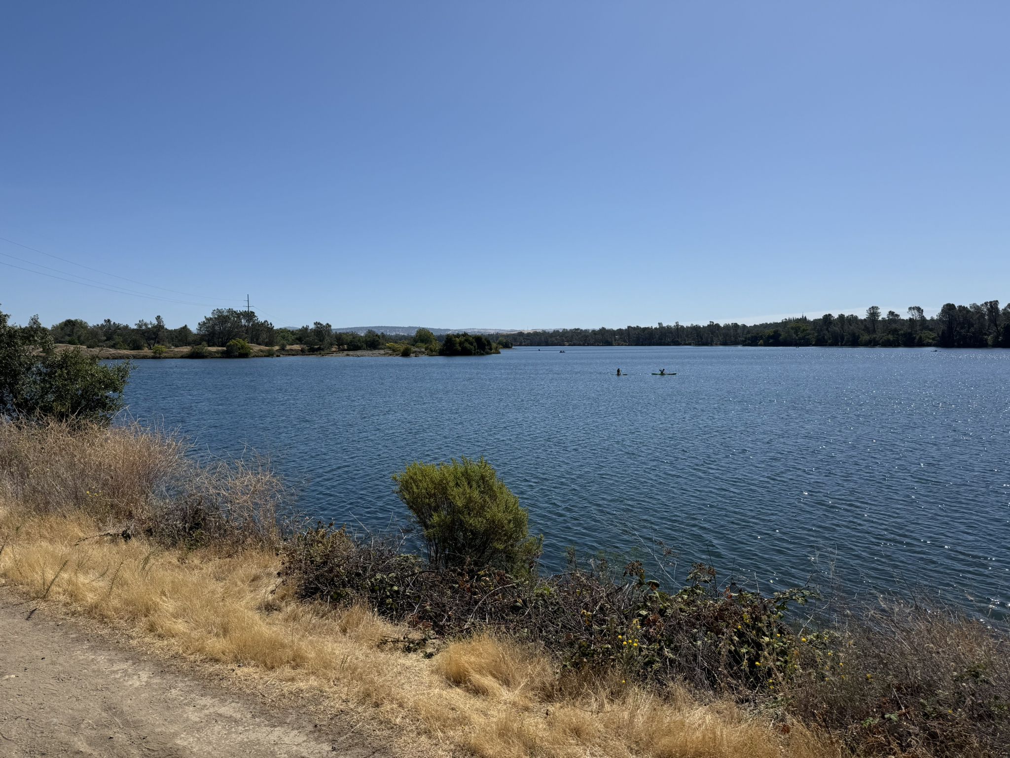 A wide blue lake with distant kayakers and a dirt path in the foreground. - American River Trail Loop