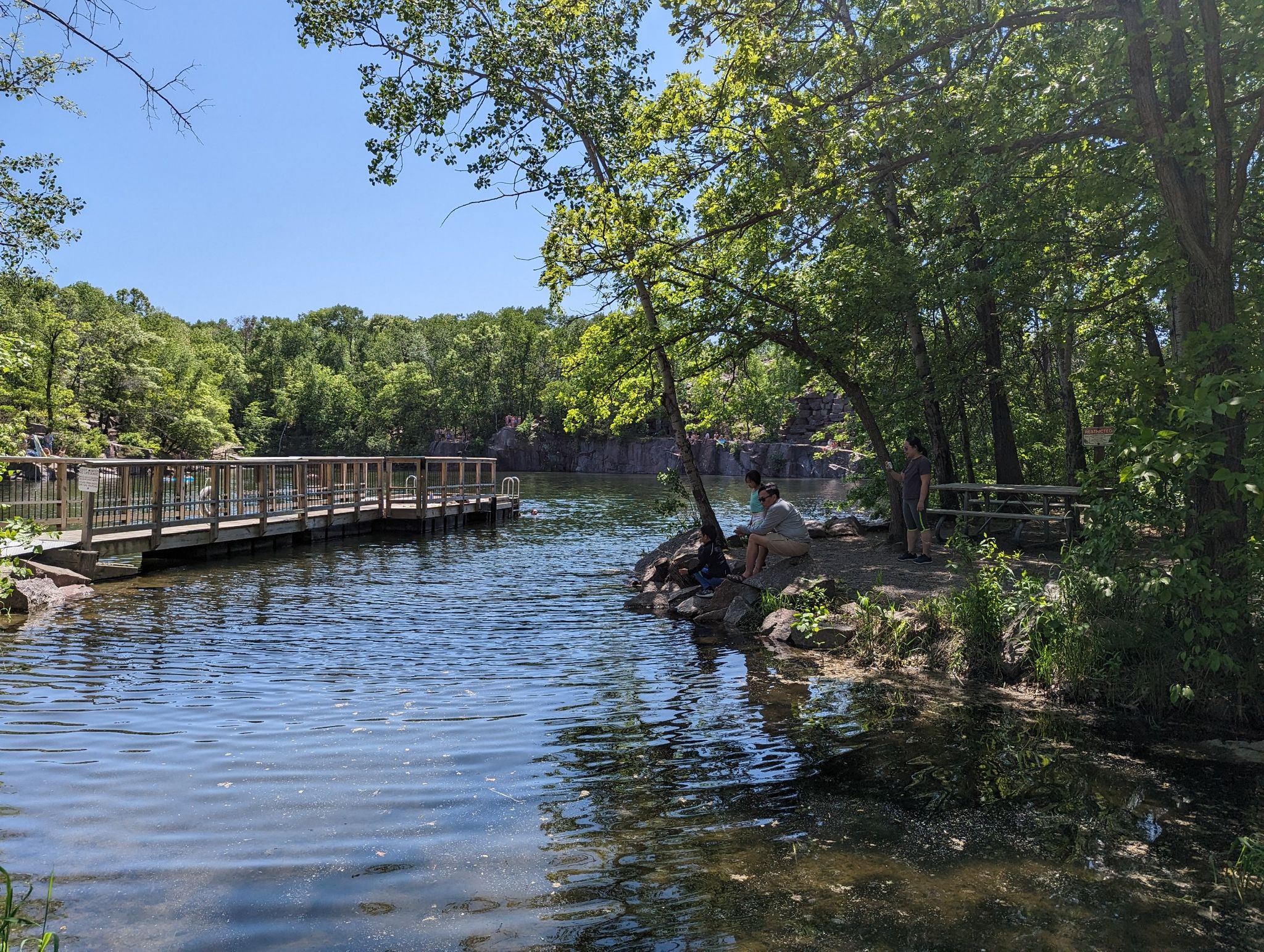 Sunny day at a lake with a wooden dock and person relaxing on the bank. - Quarry Park Outer Loop