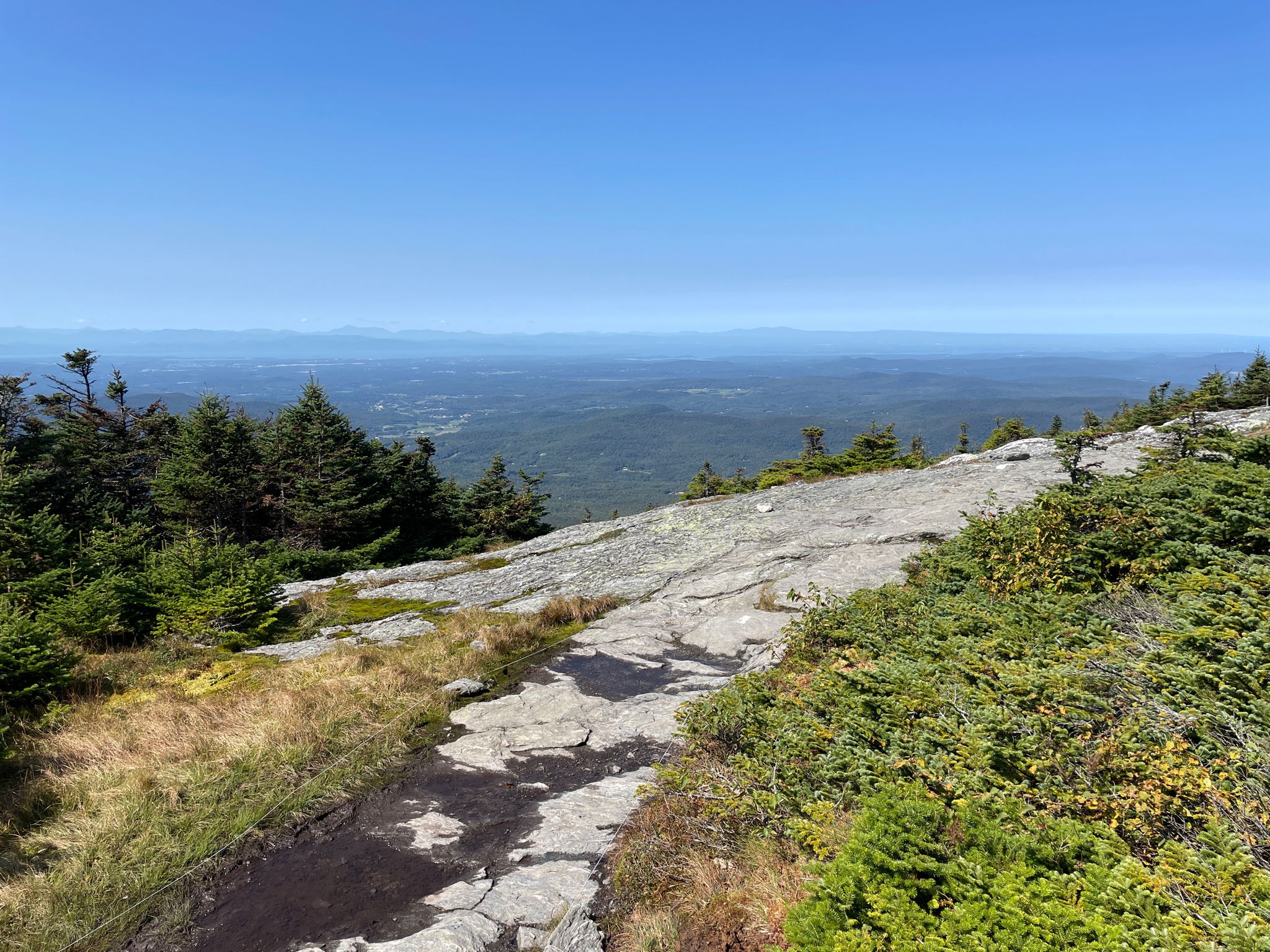 Sunny rocky mountain path overlooking a vast, hazy green valley. - Mount Mansfield Natural Area loop from Mount Mansfield Natural Area