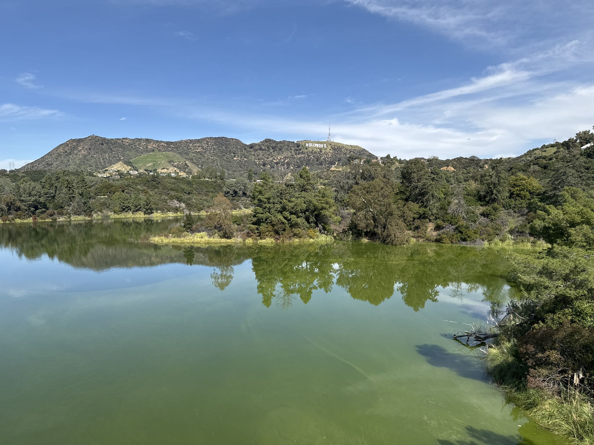 Green lake with wooded hills and distant white letters under blue sky. - Back of Hollywood Sign via Hollywood Revisor
