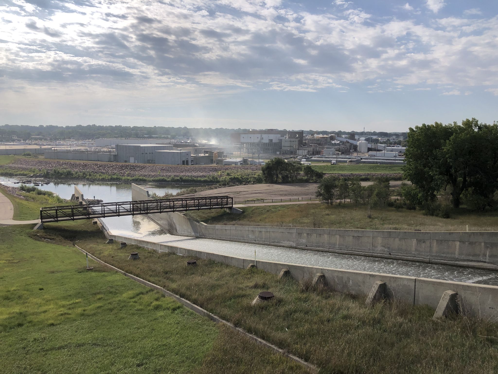 Concrete channel with flowing water, pedestrian bridge, and distant industrial buildings. - The Big Sioux River Recreation Trail and Greenway