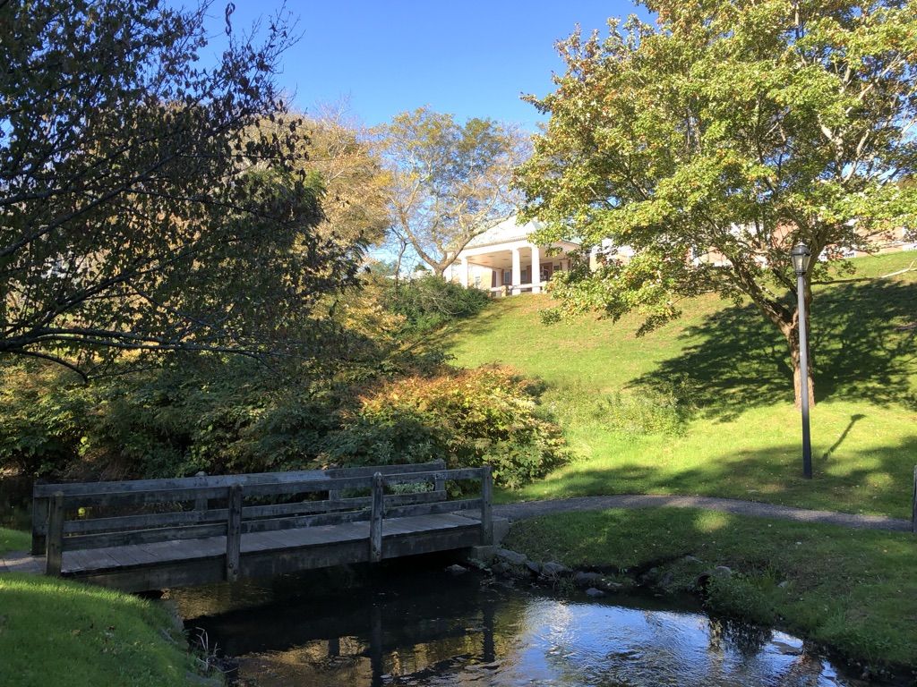 Wooden bridge over a stream, green hill with a distant white building, sunny. - Plymouth Rock – Mayflower II loop from Plymouth