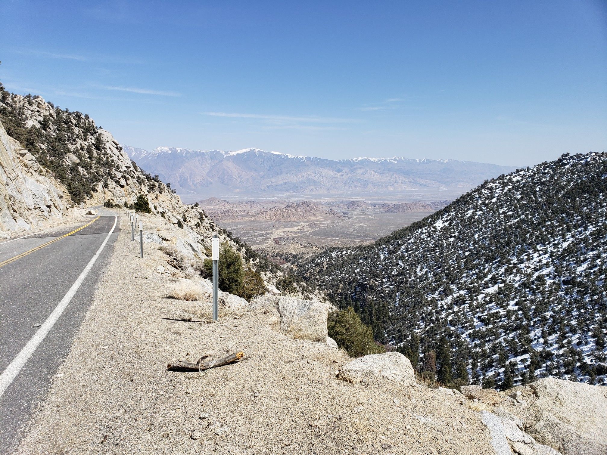 A mountain road overlooks a vast valley with distant snowy peaks under clear skies. - Whitney Portal Road