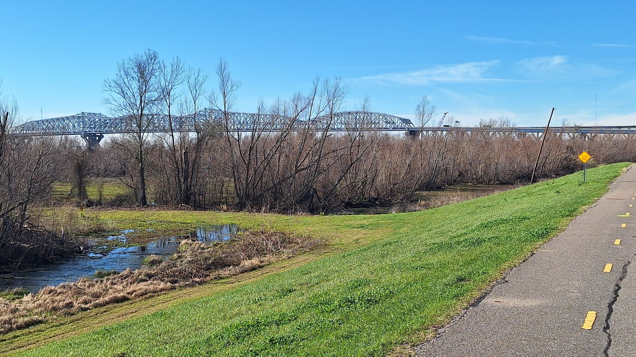 Paved path next to a river with bare trees and a distant bridge. - Mississippi River and Lake Pontchartrain Loop