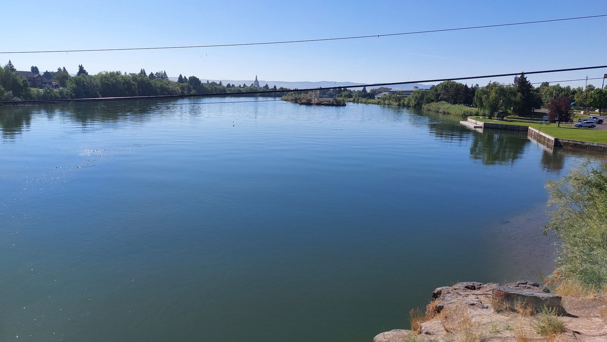 Wide, calm river reflecting a clear blue sky, with distant trees. - Friendship Garden – Idaho Falls waterfalls loop from Skyline High School