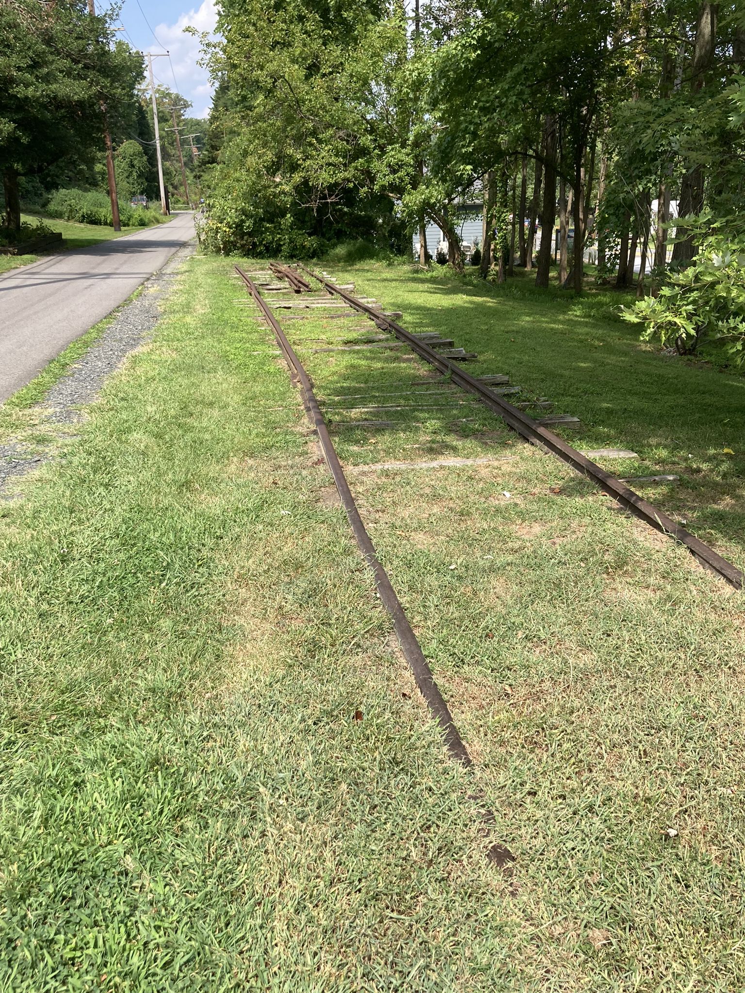 Old train tracks run alongside a paved road in bright daylight. - Baltimore & Annapolis Trail