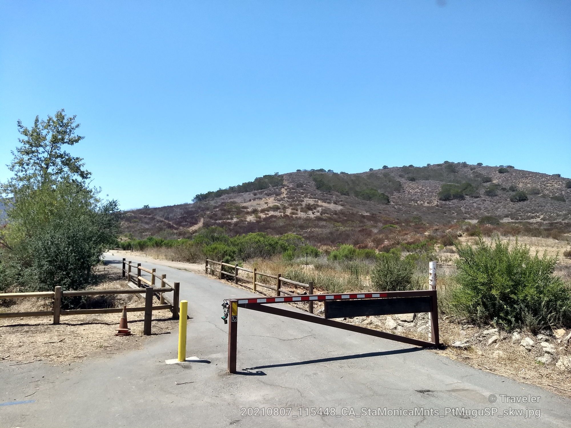 Closed gate blocks a paved road leading to dry, brushy hills. - Native Garden (trailheads) – Old Tractor loop from Dos Vientos Open Space