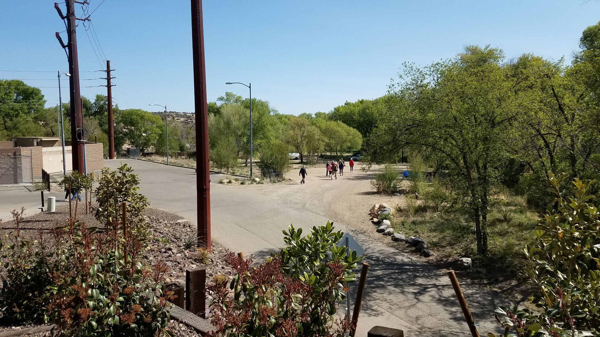 People walk on a sunny dirt path next to a road and green trees. - Montezuma Promenade