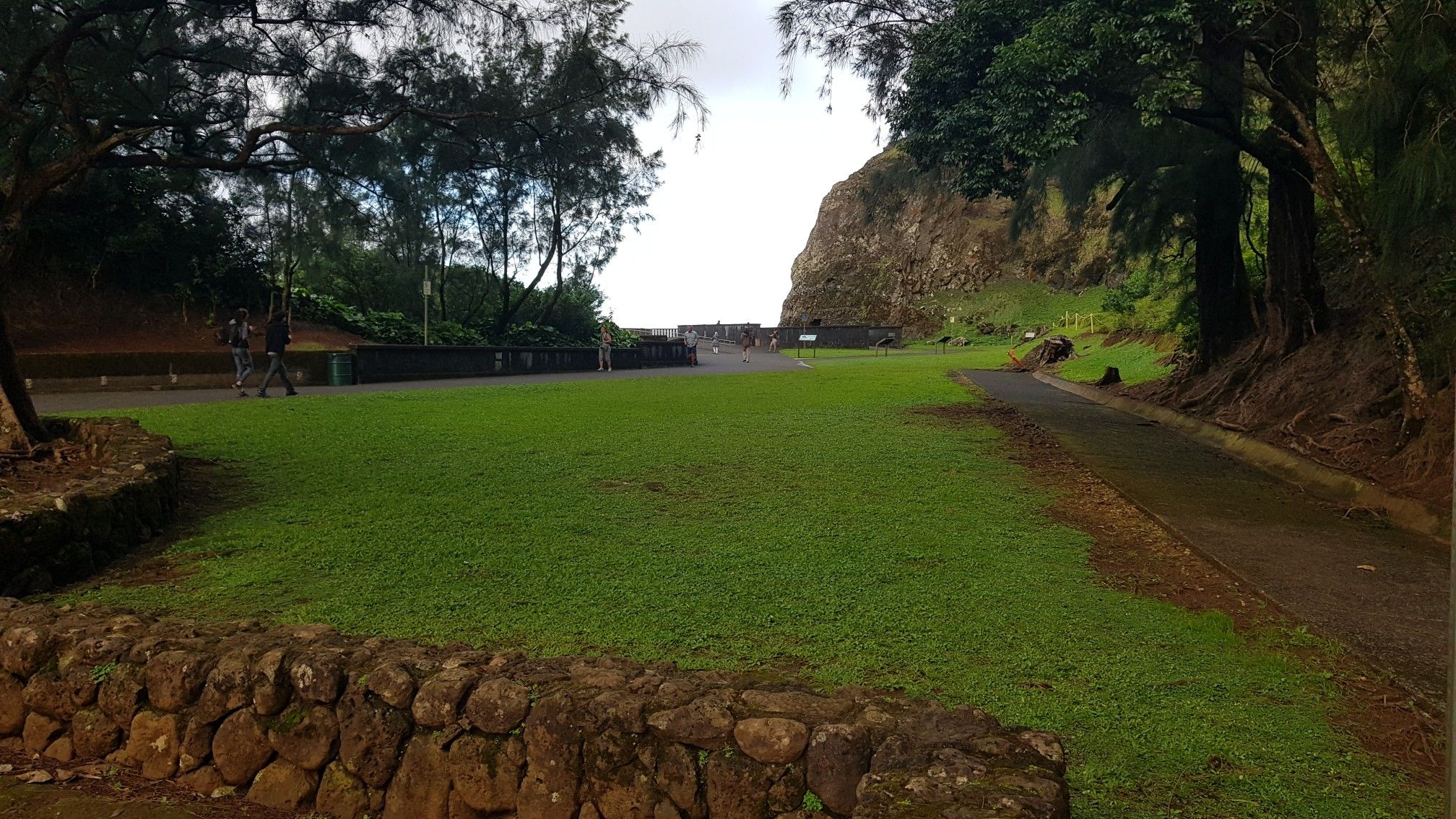 Grassy area with a stone wall, paved path, and distant rock face. - Kalihi via Kāneʻohe Bay loop