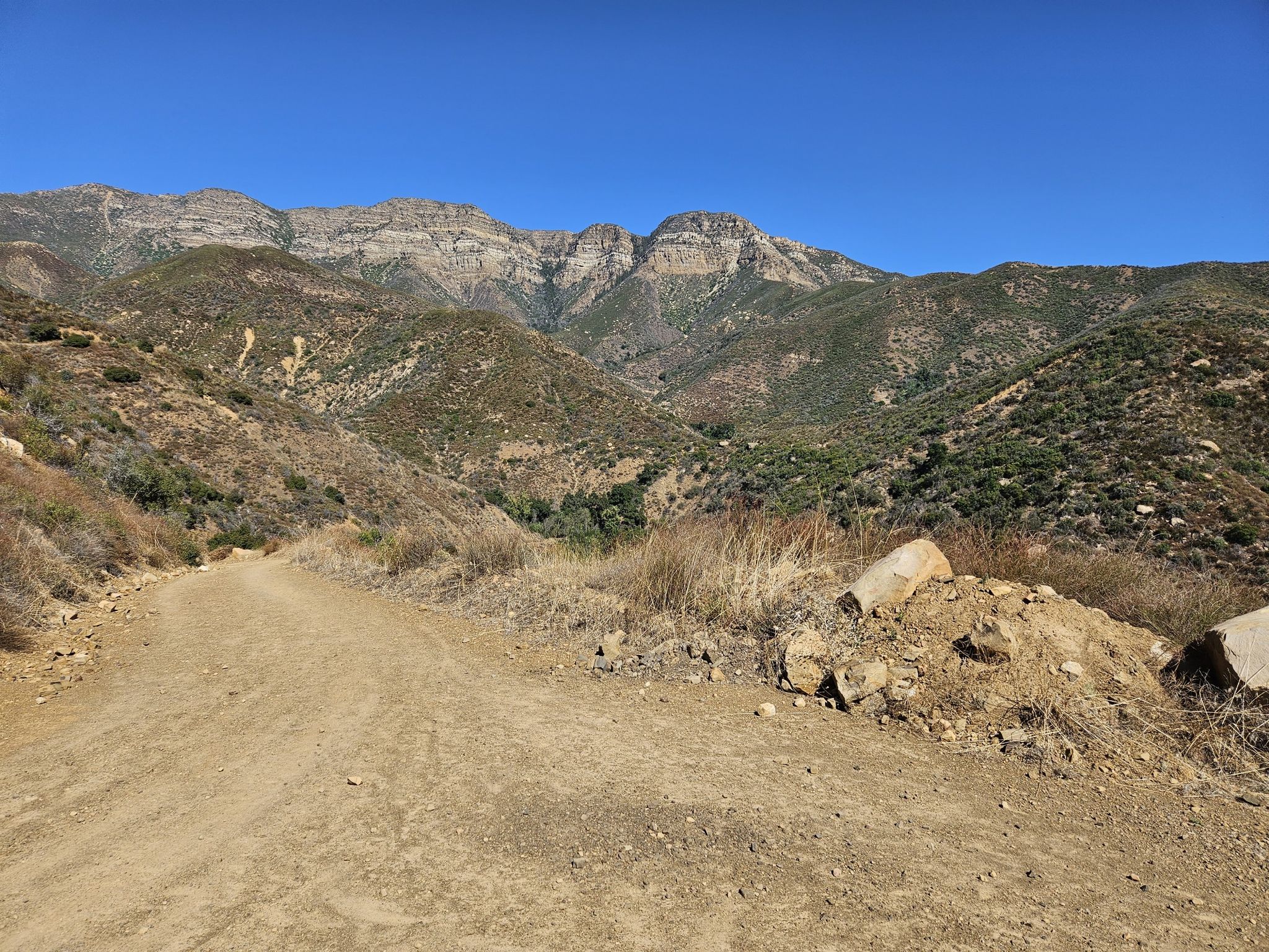 Wide dirt road with rocks leading towards distant green mountains under a blue sky. - Sisar Canyon Road – Sisar Canyon Road loop from Stagecoach Station Upper Ojai Market