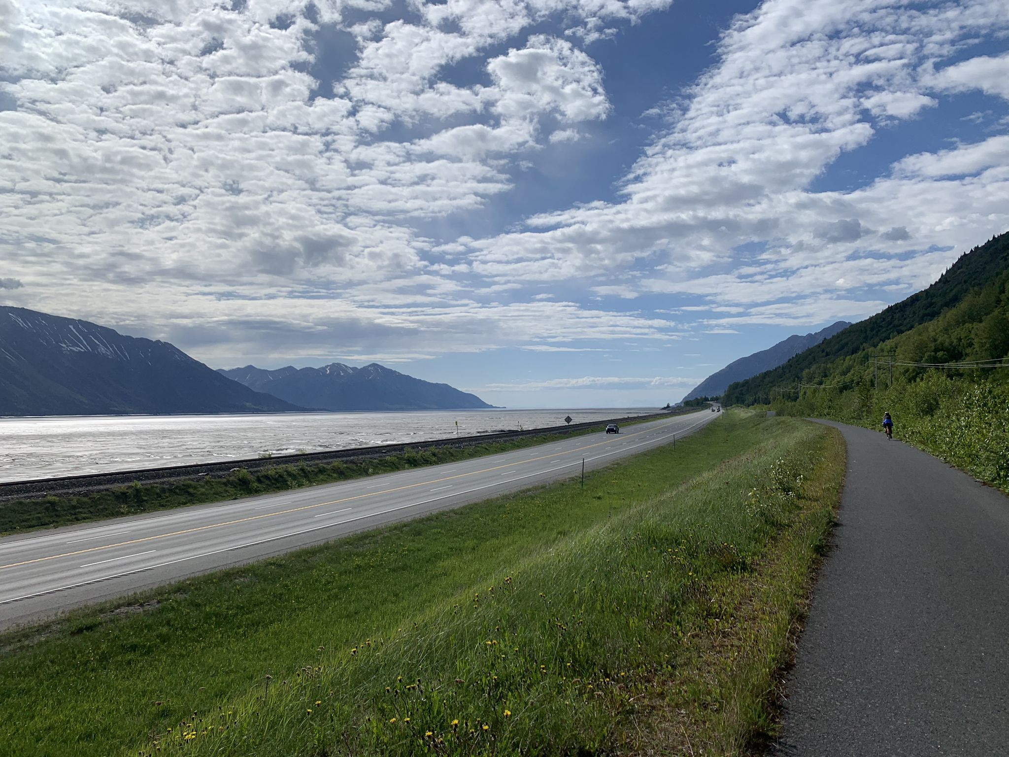 Road and paved path next to water, with mountains and a person in the distance. - Bird to Gird Pathway