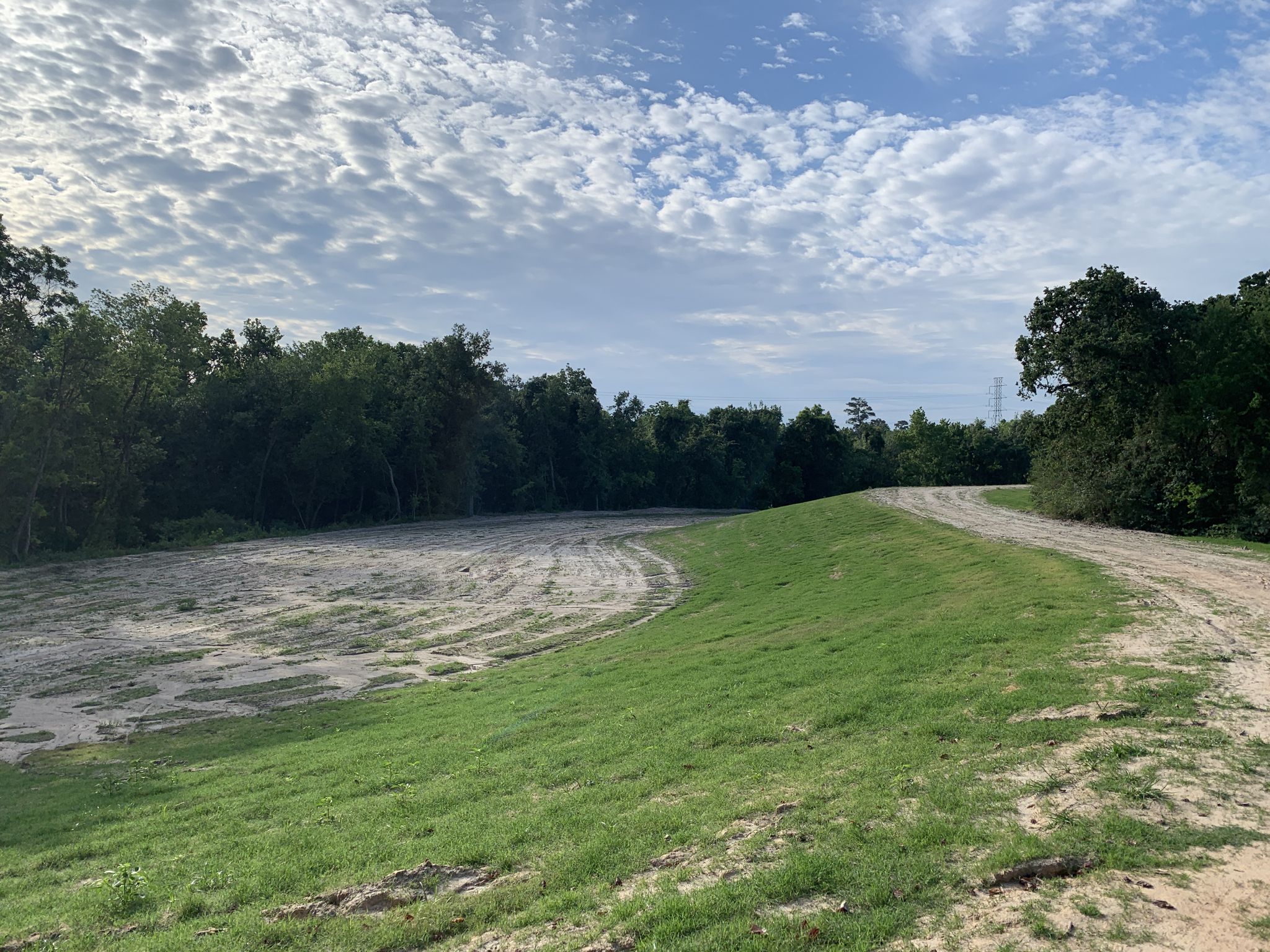 A grassy embankment curves beside a sandy field with distant trees under a cloudy sky. - Quail Trail