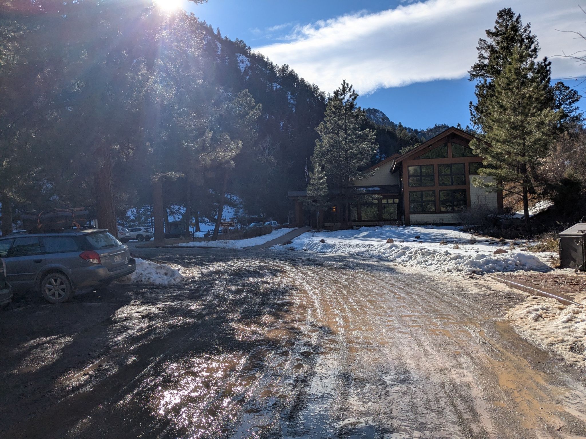 Sun shines over a snowy, muddy road leading to a building near mountains. - View of the Mountain Landscape – View of Eldorado Canyon loop from Eldorado Springs Pool