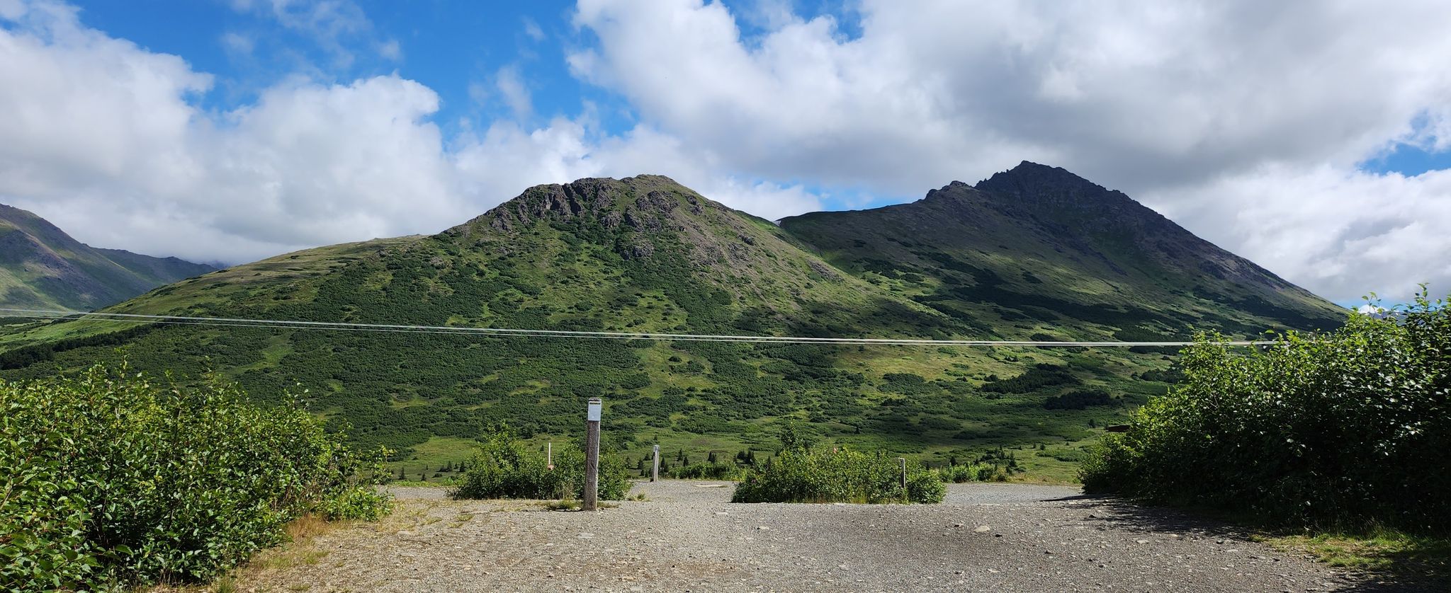 Two green, rugged mountains frame a gravel path under a bright, cloudy sky. - Powerline Pass Trail