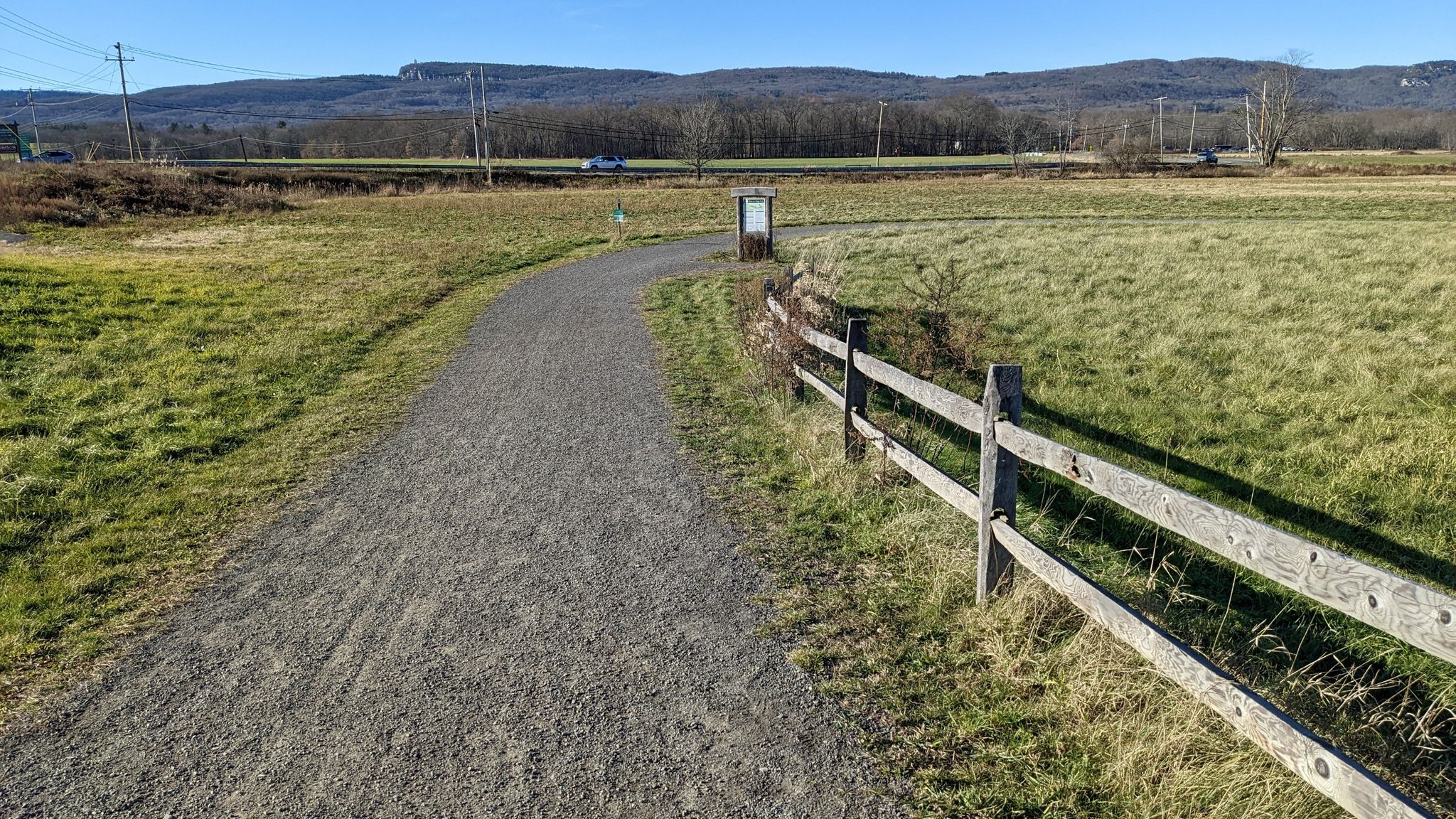Gravel path next to a wooden fence, leading through sunny fields towards hills. - Wallkill Valley Rail Trail Loop
