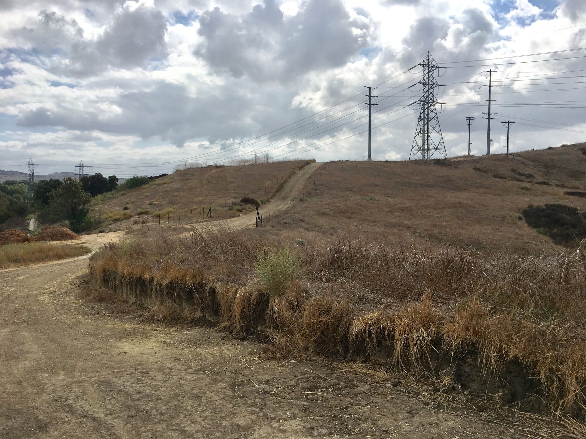 Dirt path winding up a dry, grassy hill with power lines under a cloudy sky. - Waterworks Flow Trail – Trabuco Creek trails loop from Los Patrones